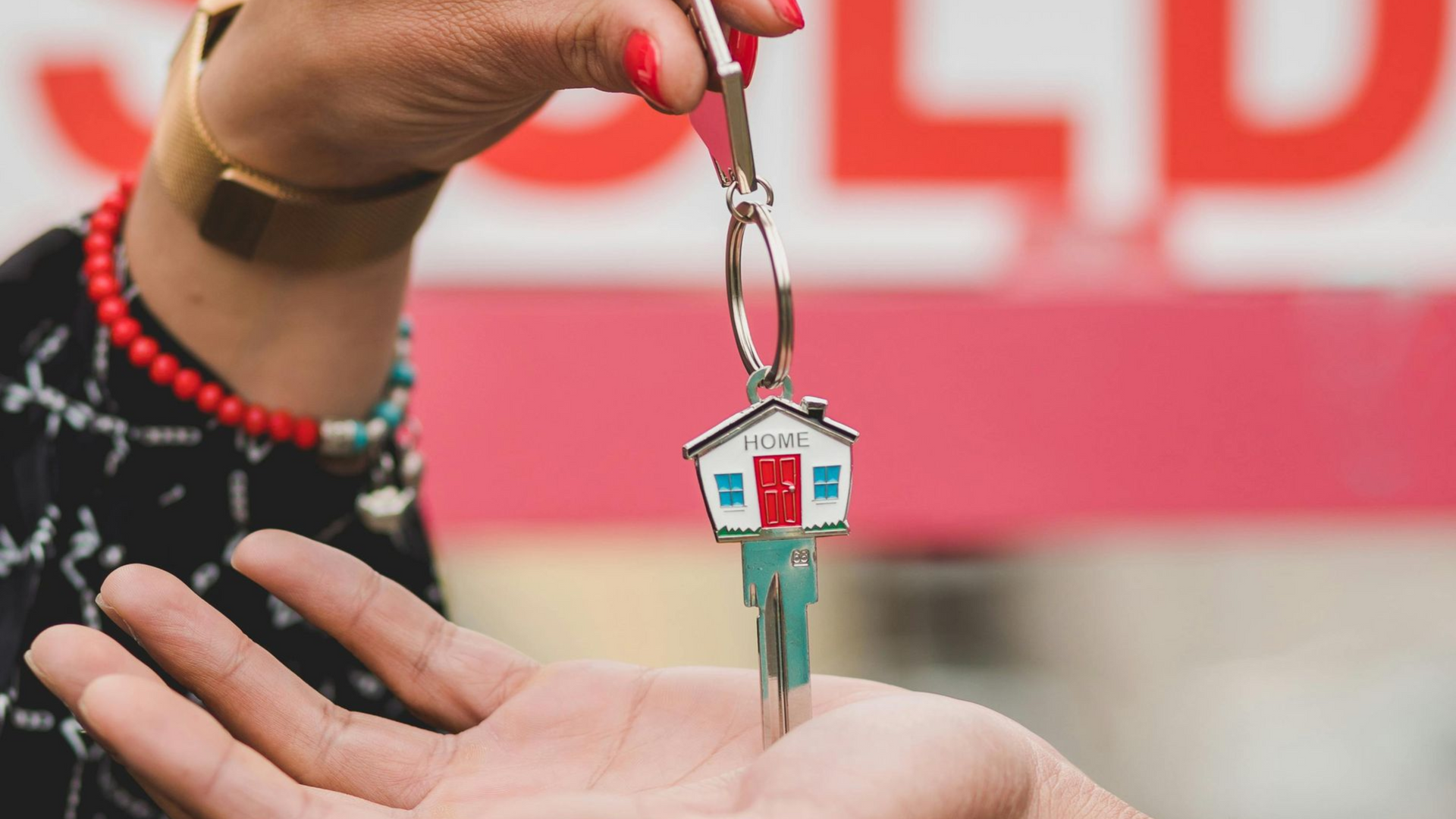 Hand giving house key with house-shaped keychain in front of a “SOLD” sign.