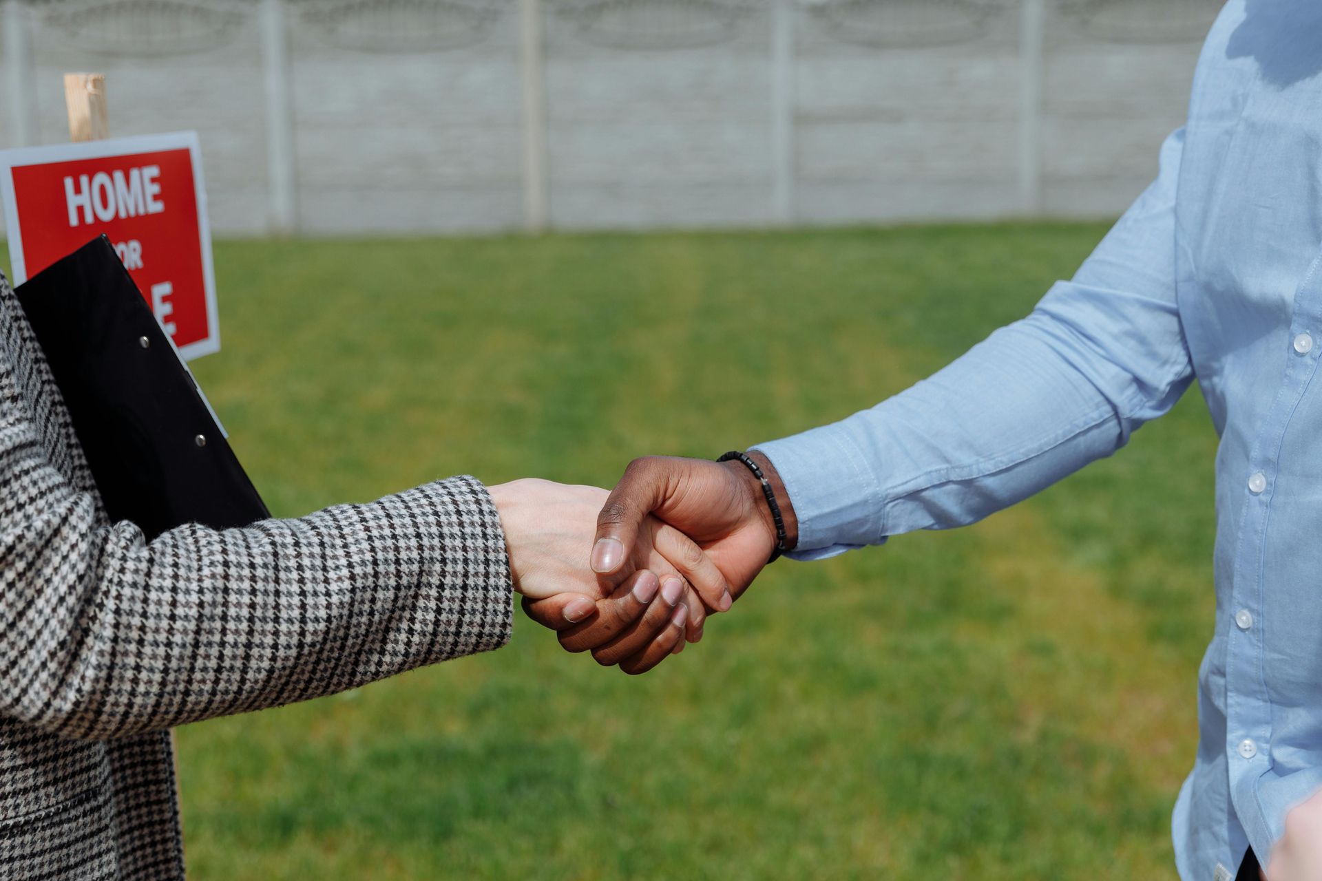 Two people shaking hands in front of a house for sale sign