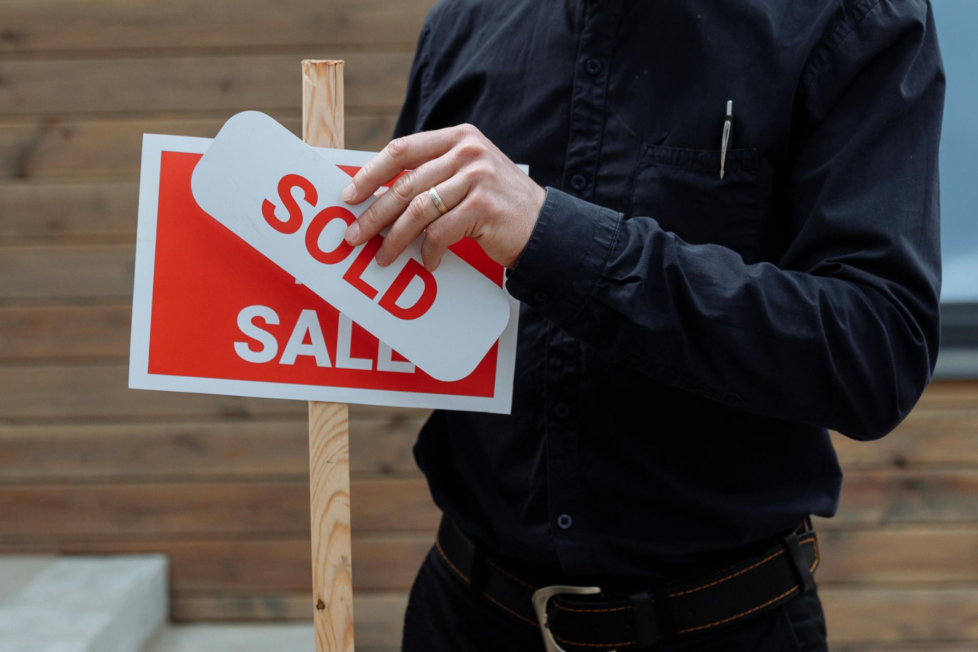 Person holding a red and white “SOLD” sign on a wooden post outdoors