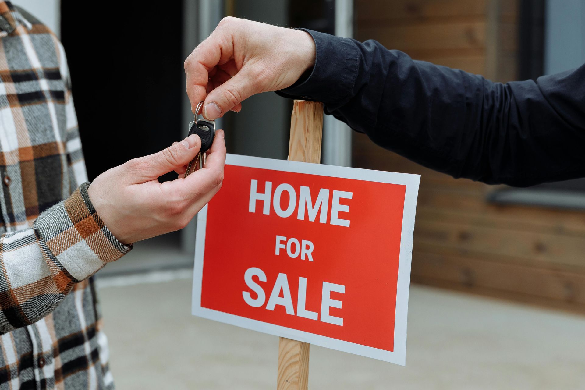 Hands exchanging house keys beside a red “HOME FOR SALE” sign outdoors