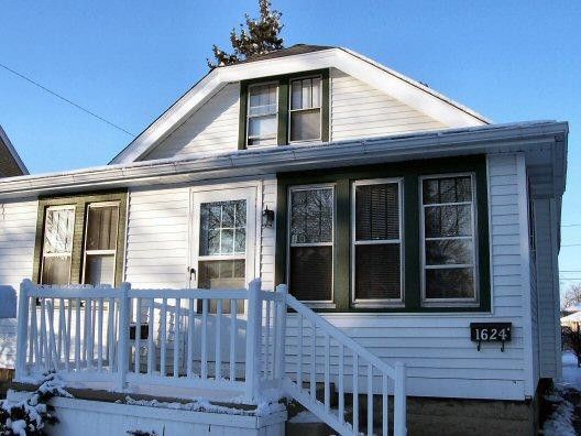 A small, white, one-story house with a white front porch railing and green-trimmed windows under a clear blue sky.
