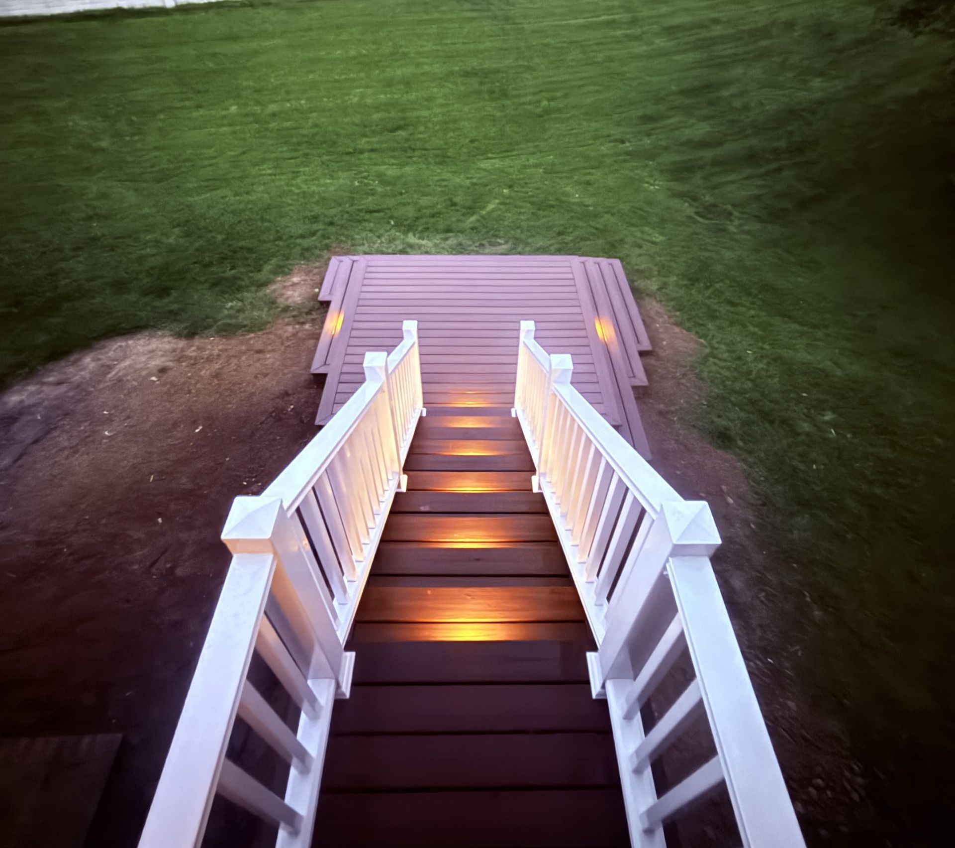 Lit white-railed stairs with glowing step lights descending to a wooden deck over grass