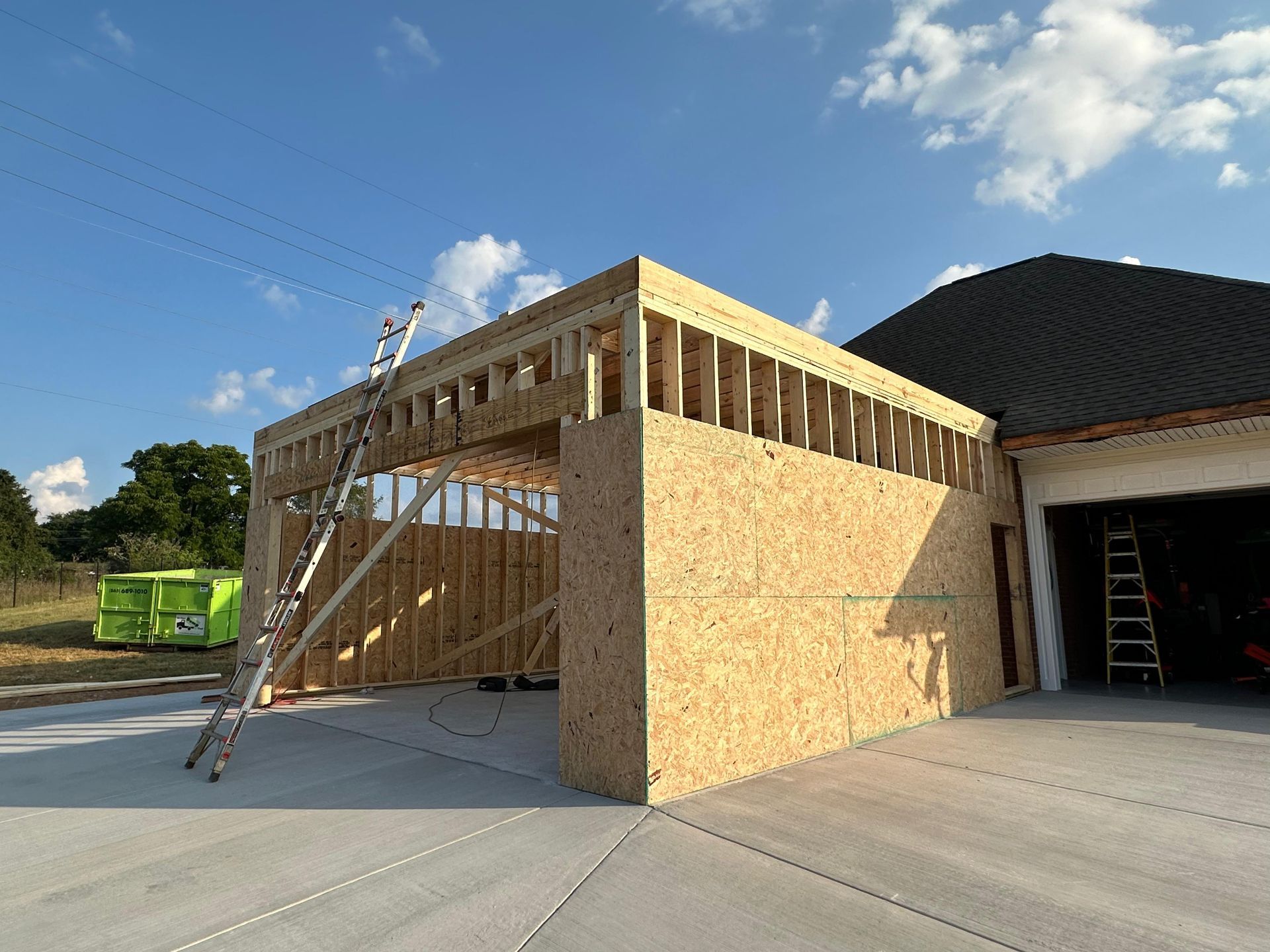 Partially framed garage addition with OSB sheathing and ladders on a driveway under a blue sky