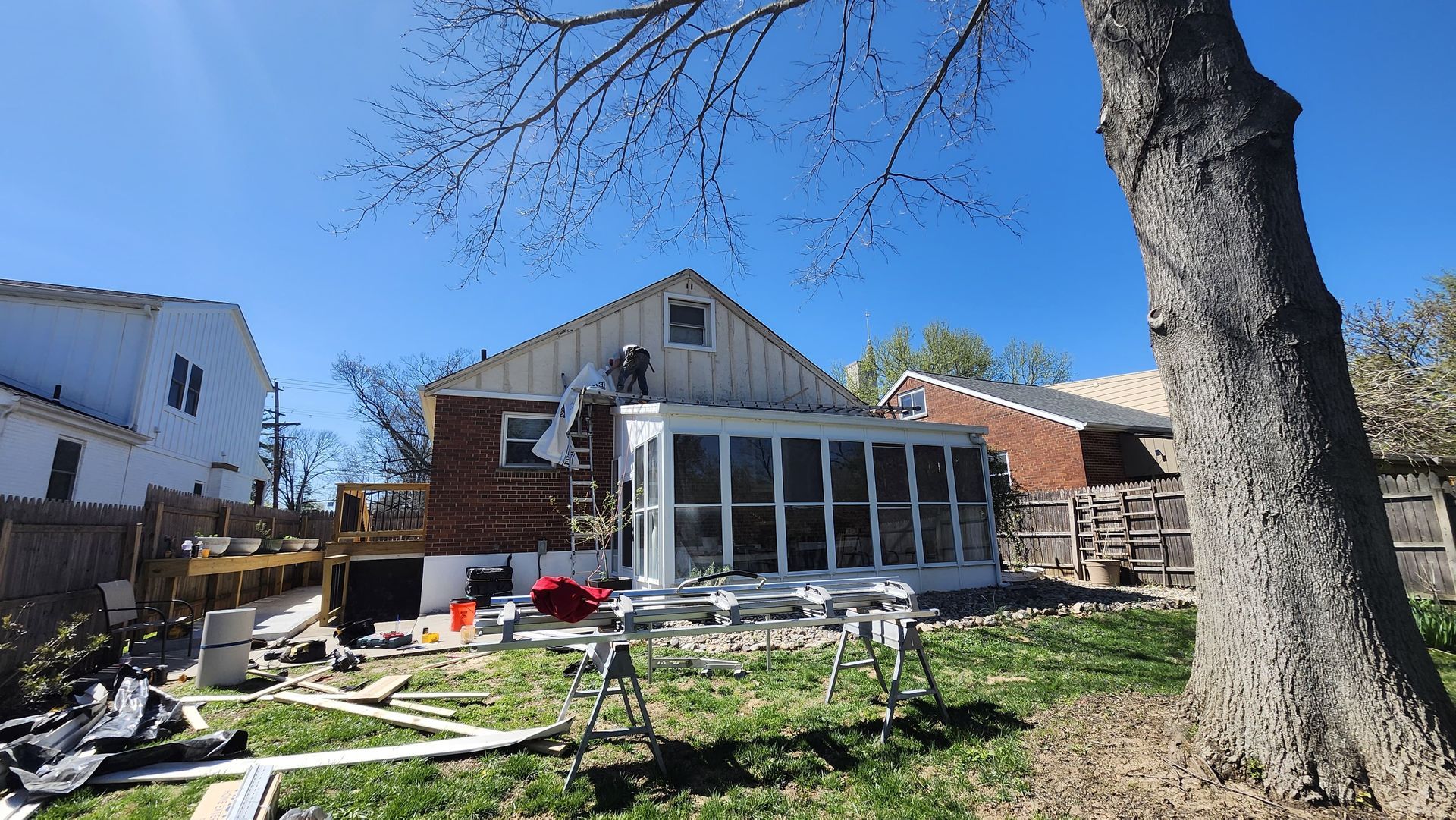 Backyard with a small house, sunroom, large tree, and scattered construction materials under a blue sky