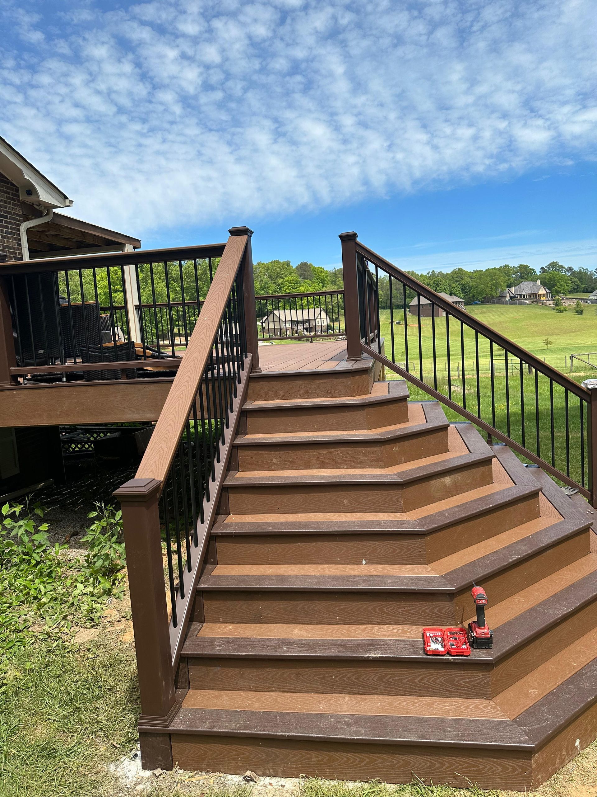 Outdoor wooden staircase with brown railings beside a porch, leading down to a grassy yard under a blue sky.