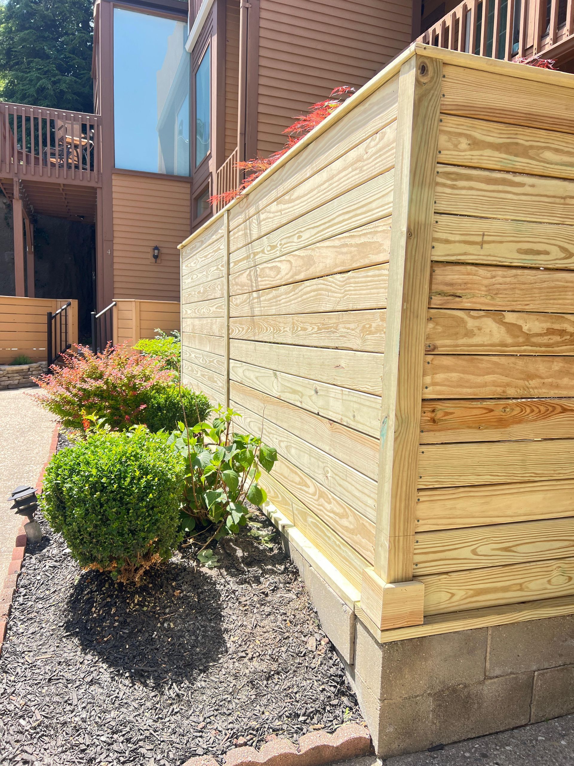 Wooden privacy fence beside a gravel bed with shrubs and a brick house in bright sunlight
