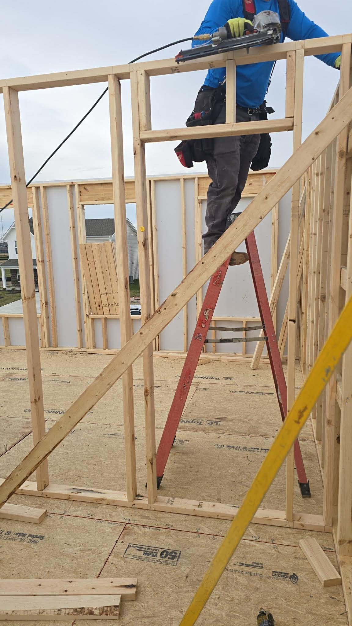 Two workers on a wooden frame wall, one on a ladder at a construction site under clear sky