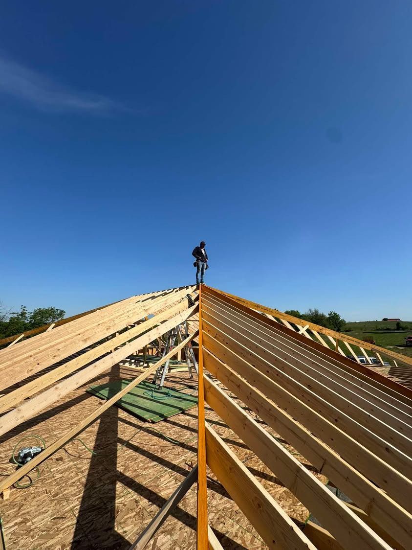 Roof framing under construction with a worker standing on the ridge against a clear blue sky