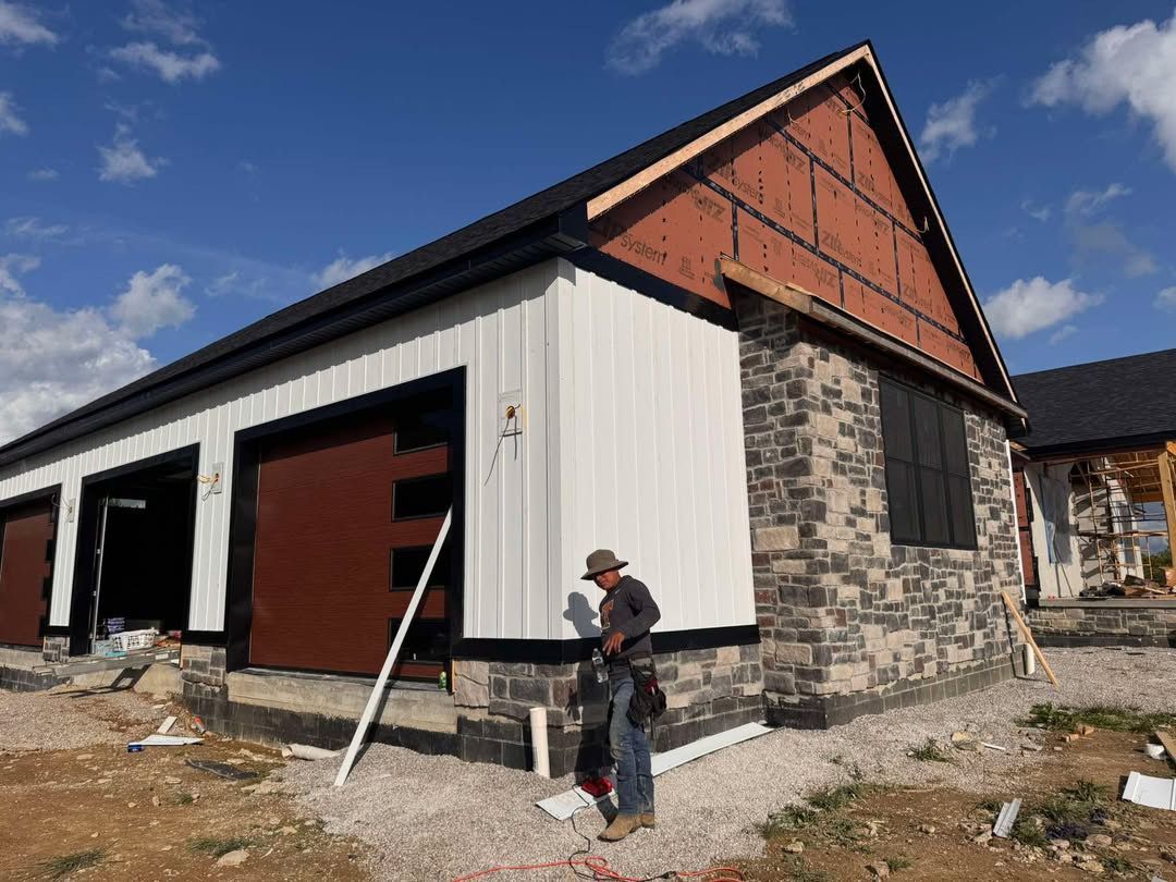 Partially built house with stone and white siding; a worker stands outside under a blue sky.