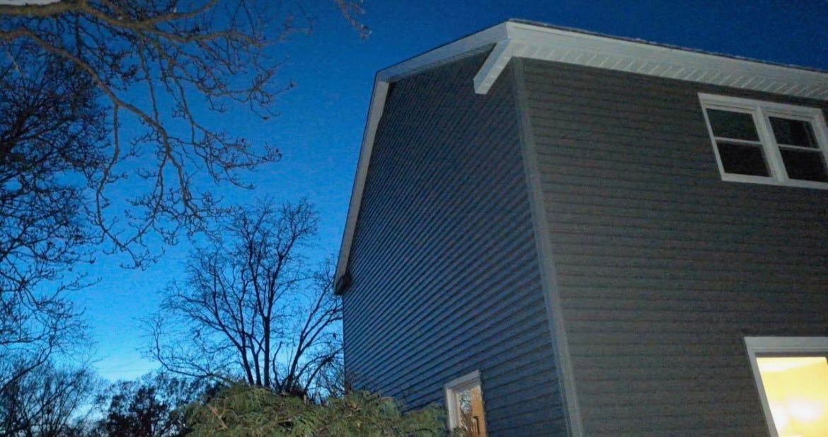 Blue evening sky behind a gray house with bare tree branches silhouetted on the left.