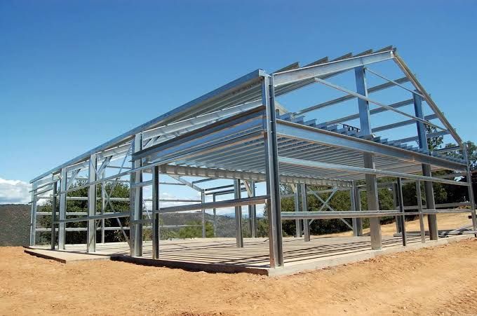 Steel frame of a large building under construction on a dirt lot, with clear blue sky behind it