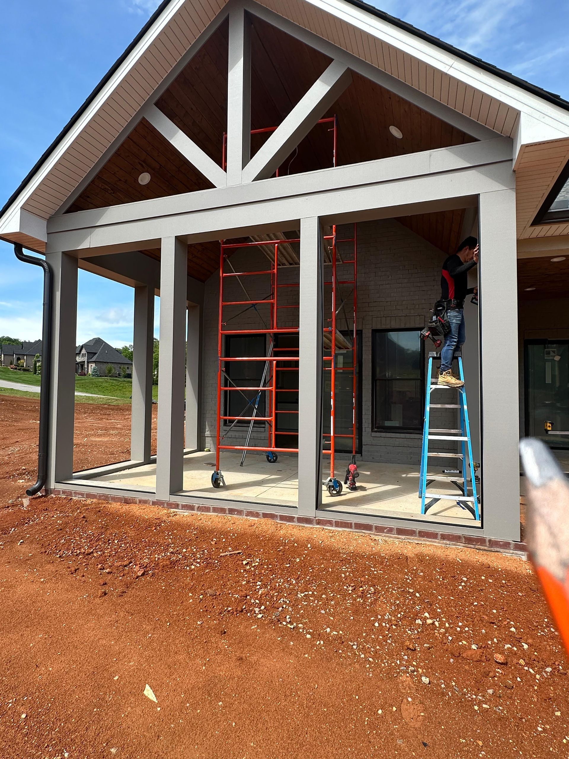Partially built house with front porch and workers' ladders, framed against a red dirt lot