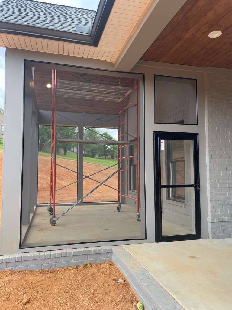 Construction scaffold in front of a large window beside a black door on a house exterior.