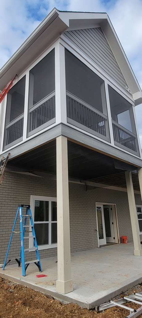 Two-story house under construction with screened porch, blue ladder, and unfinished concrete foundation