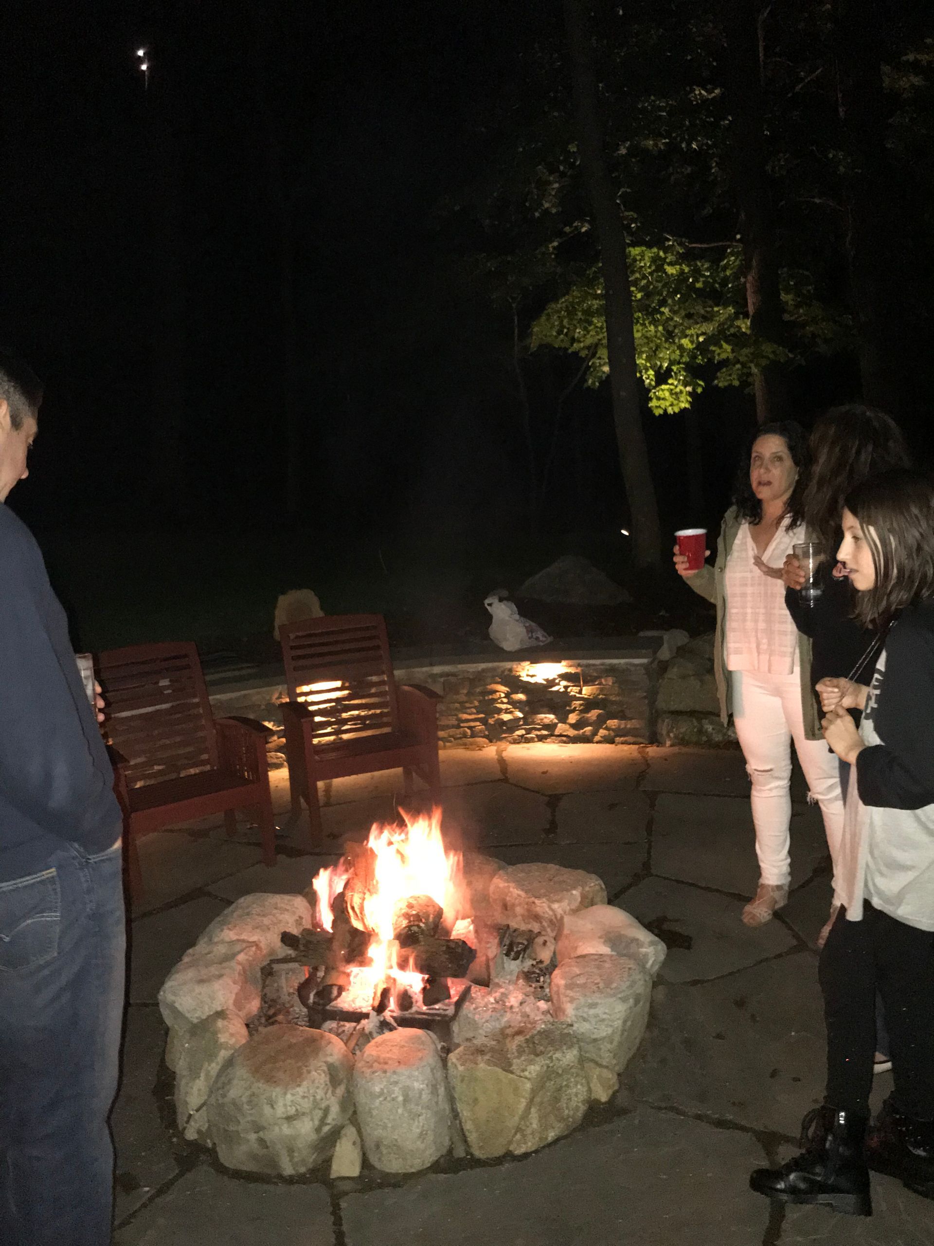 People standing around a small outdoor fire pit at night, with chairs and a red cup nearby.