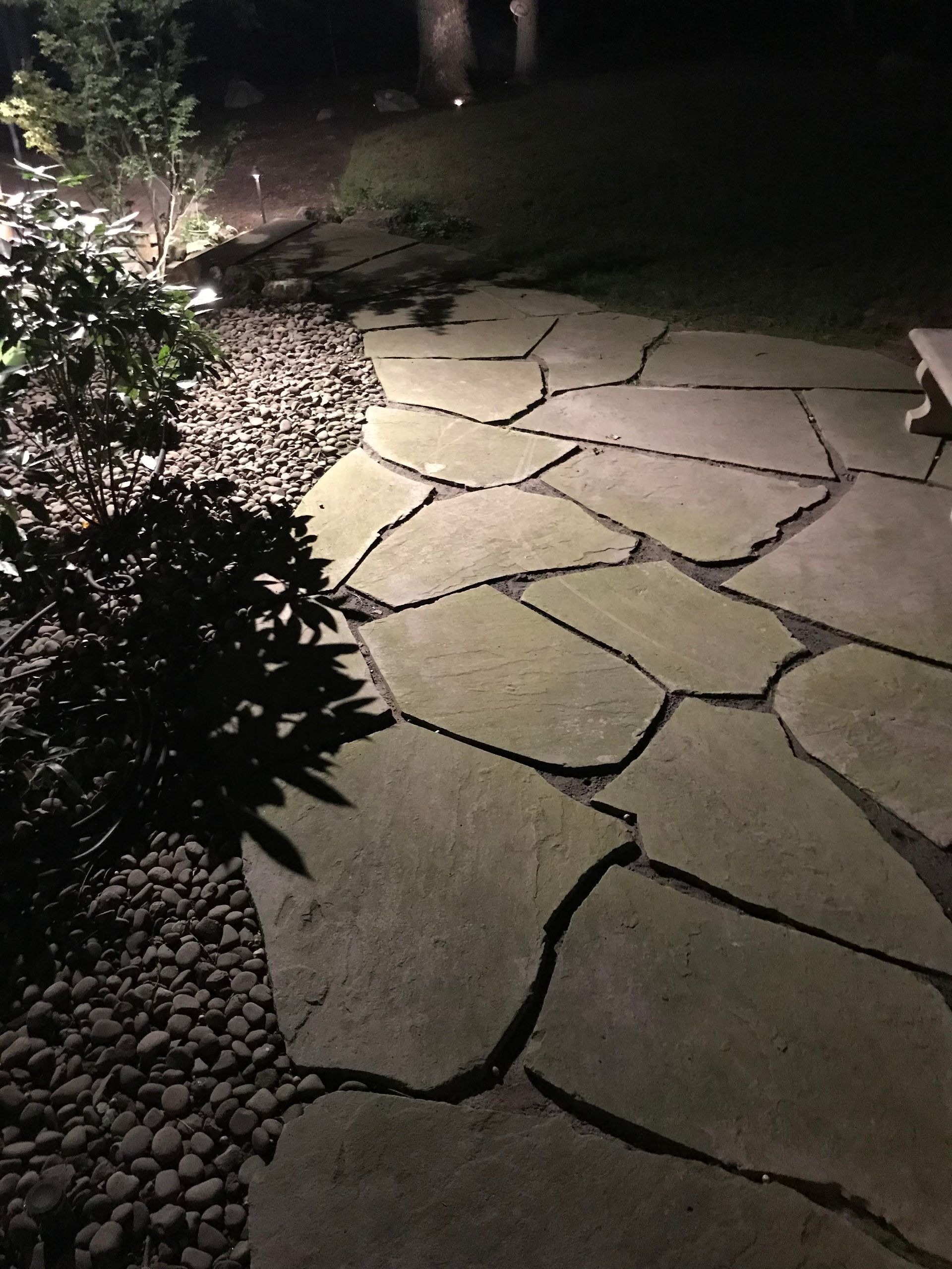 Stone garden path at night, lit by warm lights beside dark plants and pebbles.