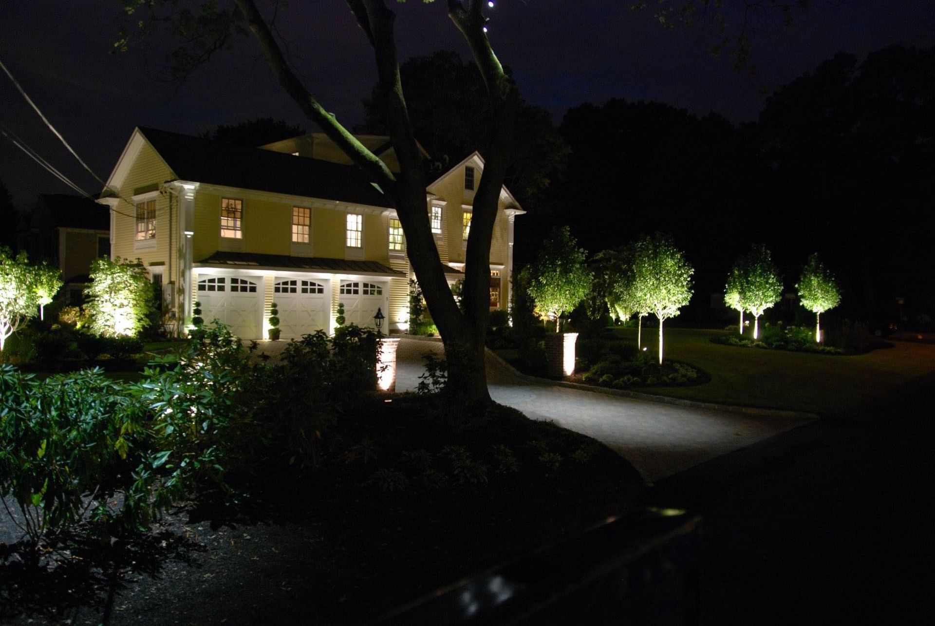 Nighttime view of a lit house and driveway, with trees and landscaped shrubs in the foreground.