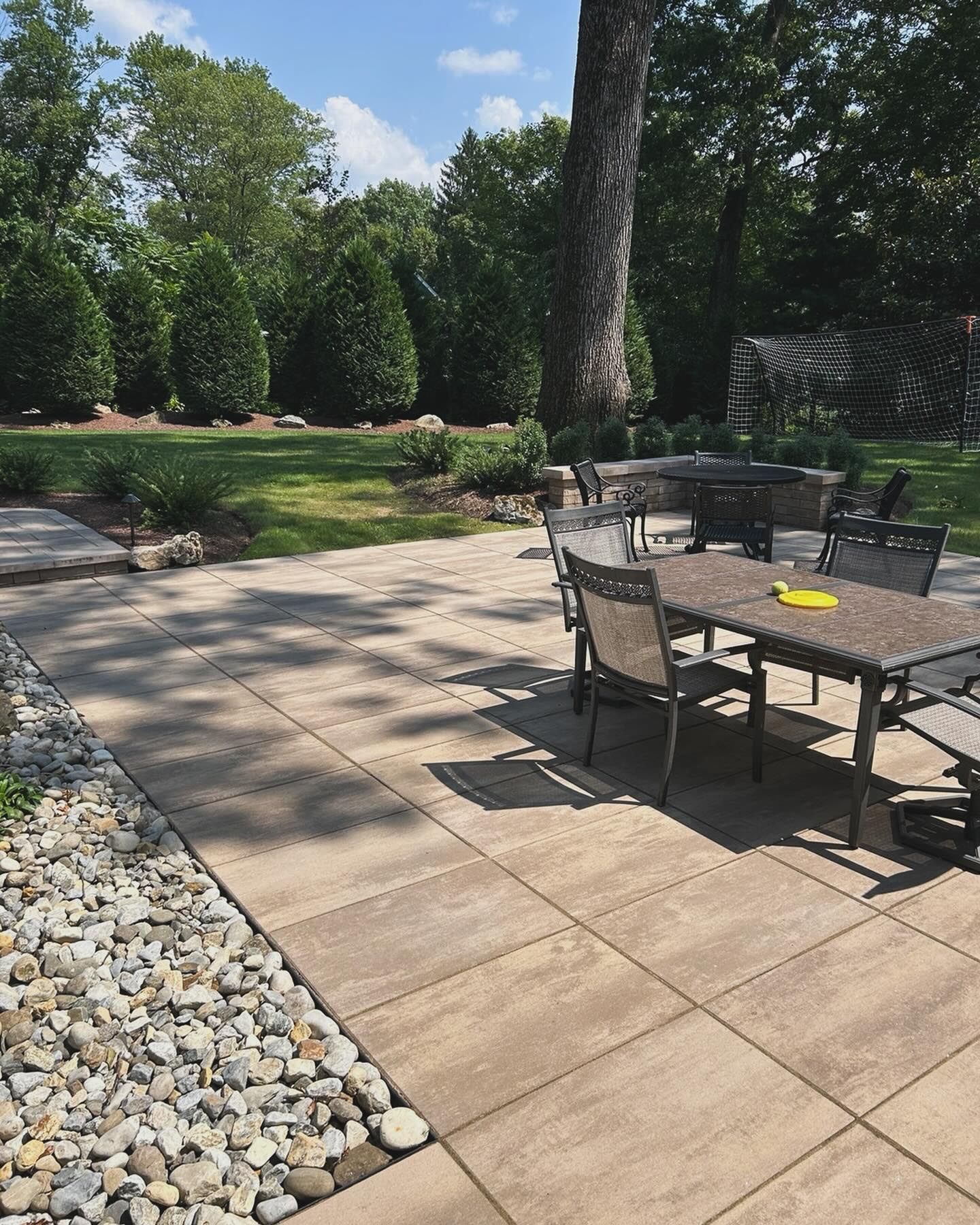 Patio with outdoor dining table and chairs beside a landscaped lawn and trees on a sunny day