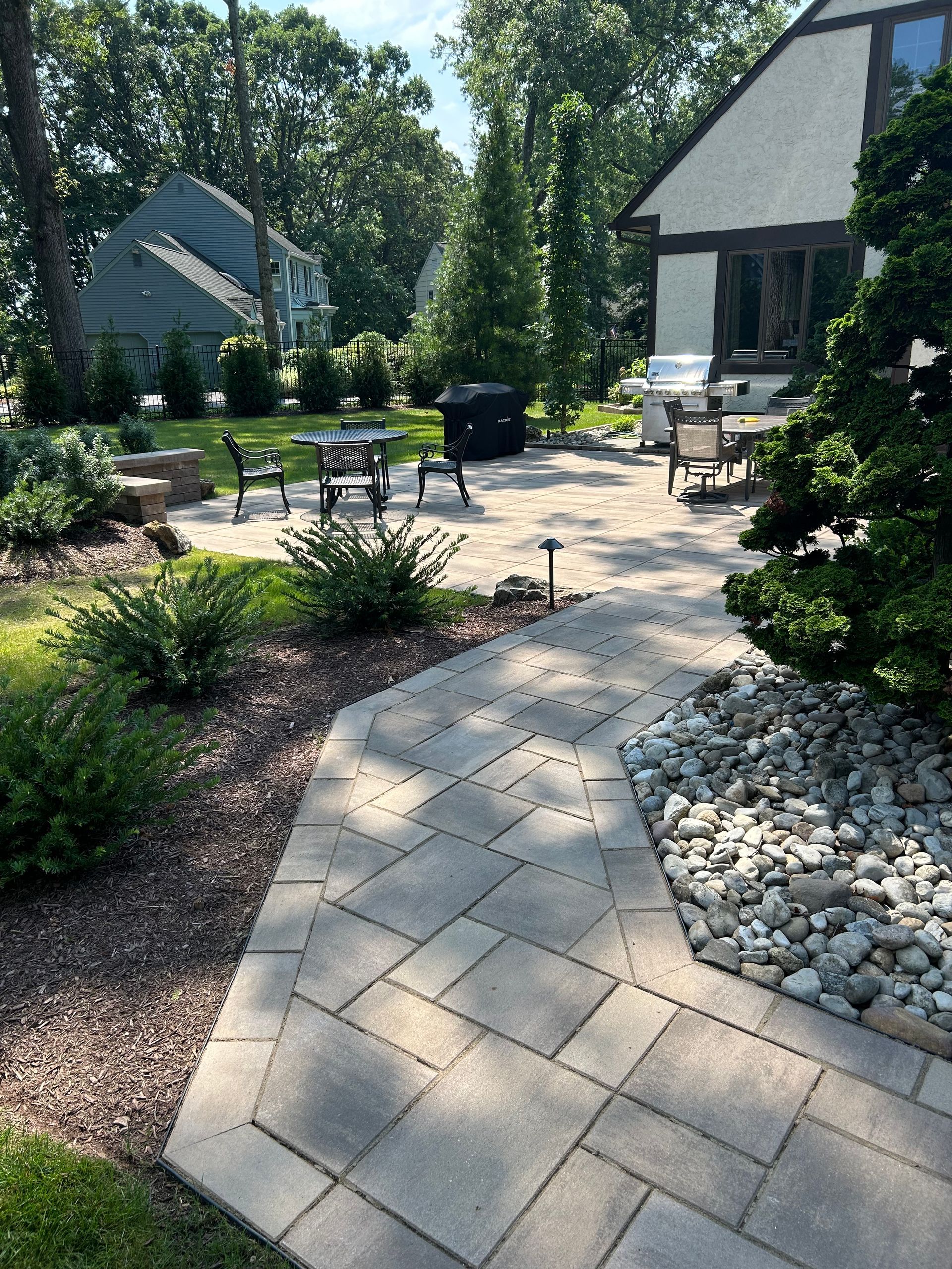 Curved stone walkway leading to a sunny patio with tables, chairs, and landscaped garden beds.