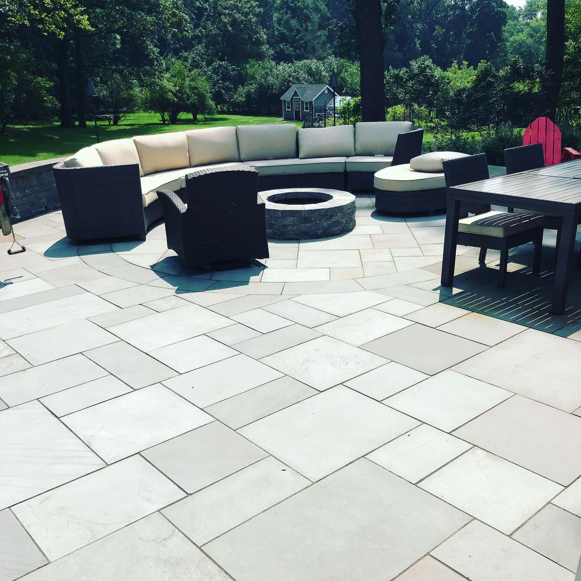 Patio with light stone pavers, curved outdoor sofa, fire pit, and dining table surrounded by trees