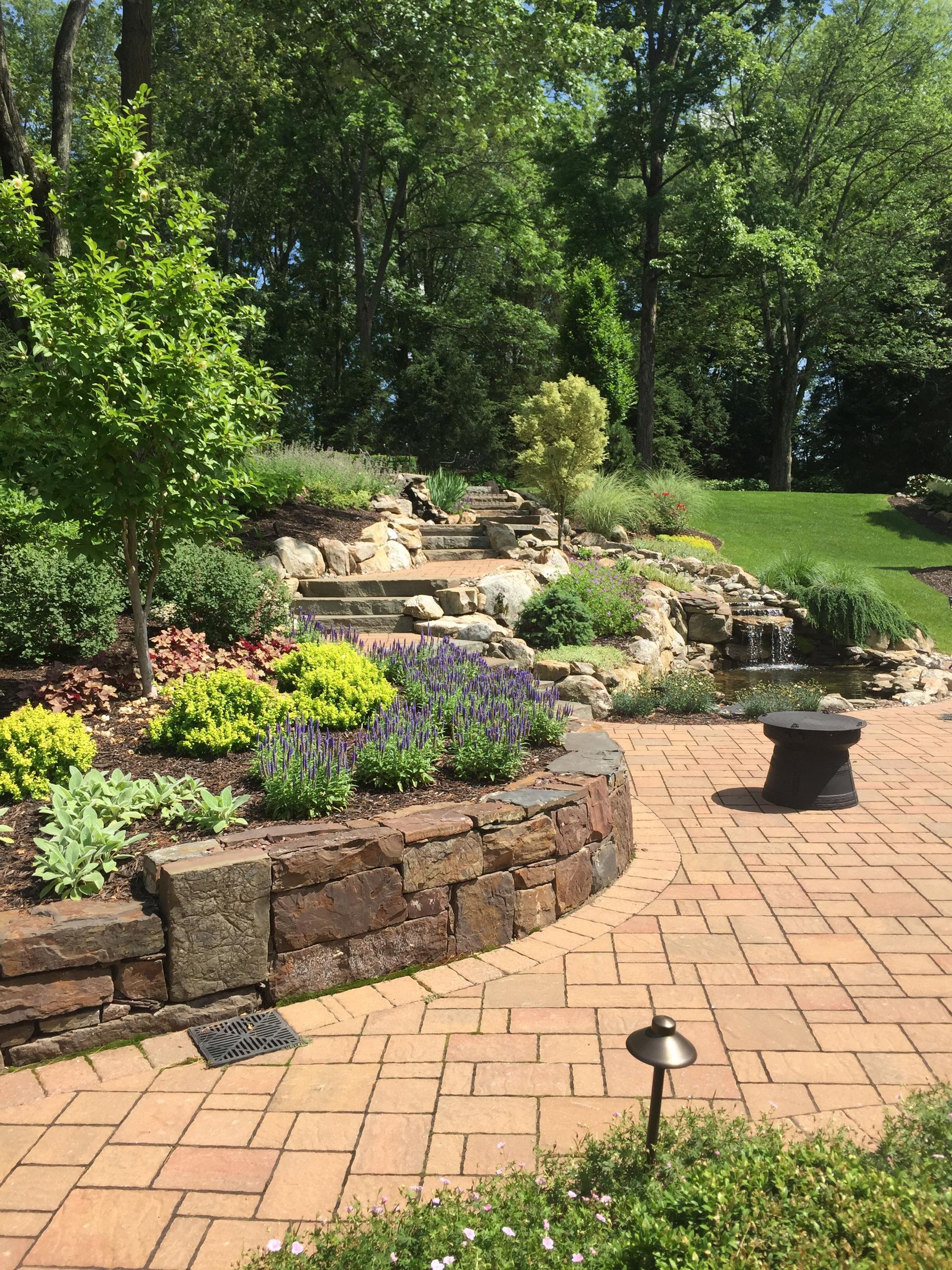 Brick garden path beside stone retaining wall, purple flowers, and lush green trees in a sunny backyard.