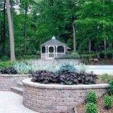 Small white gazebo in a wooded park behind a circular stone planter.