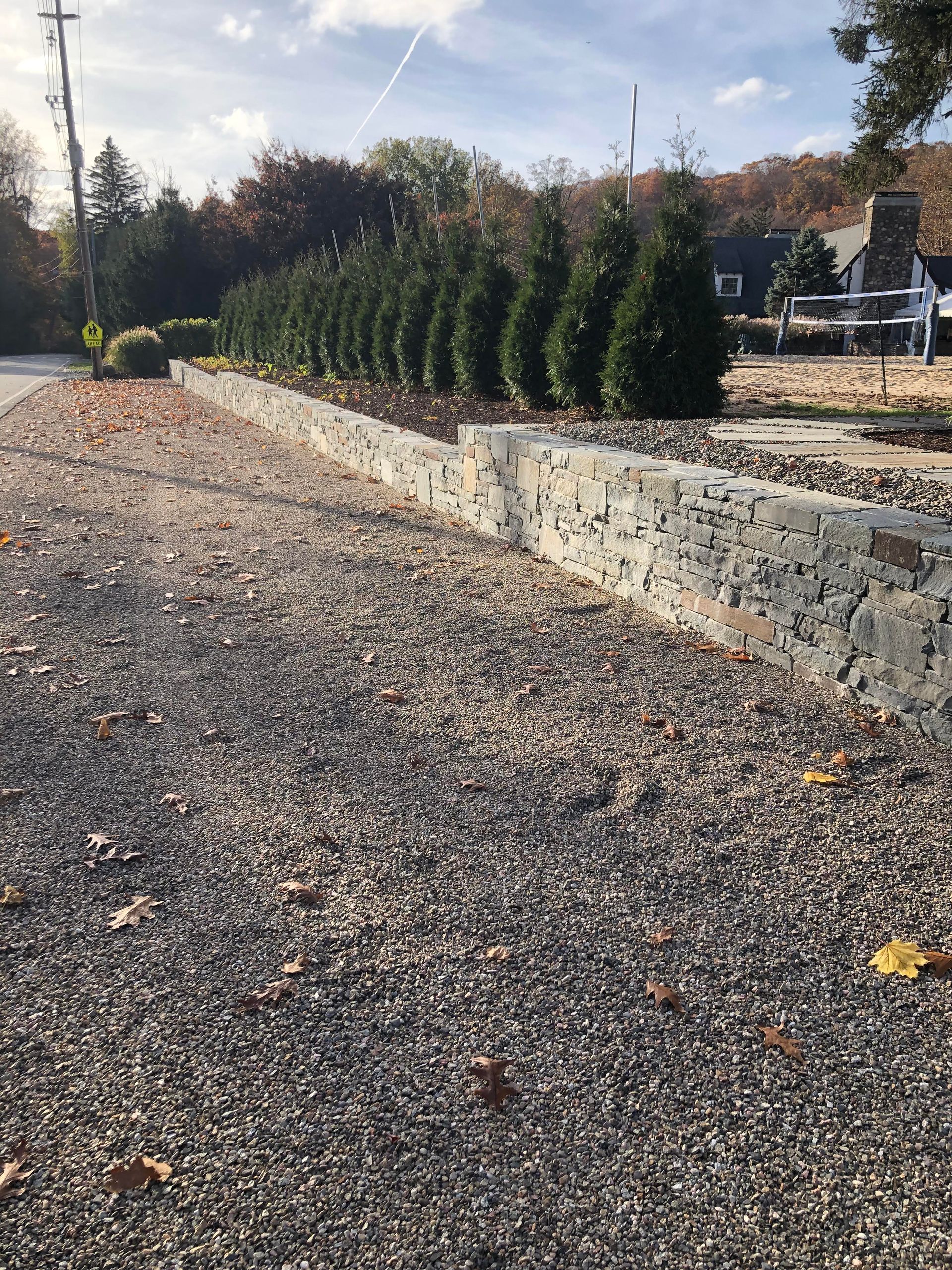 Gravel driveway beside a low stone wall and tall evergreen hedges under a partly cloudy sky