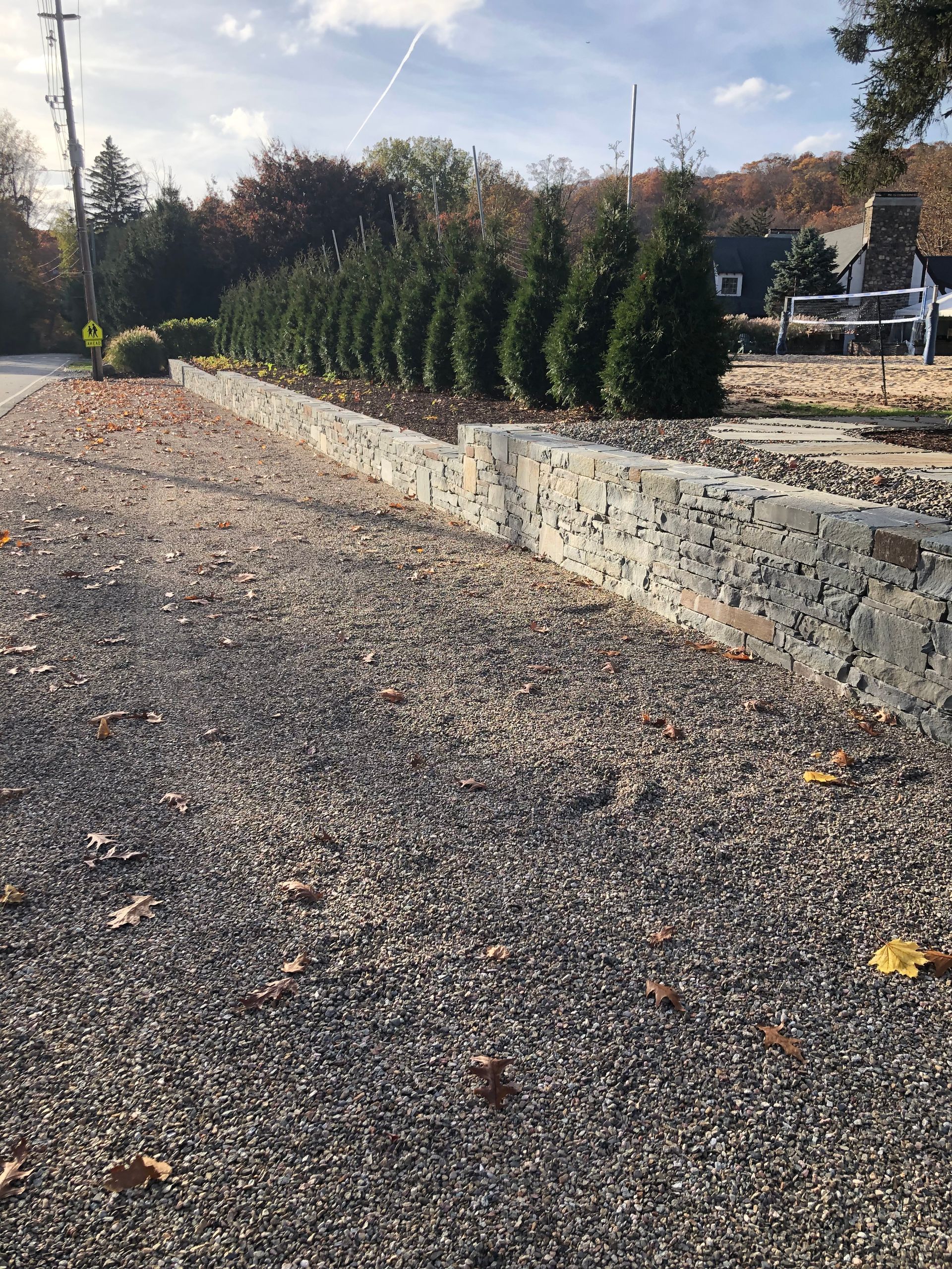 Gravel driveway beside a stone retaining wall and trimmed evergreen hedge on a sunny day
