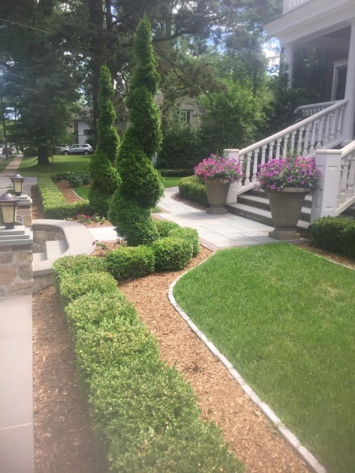 Front yard garden with trimmed shrubs, brick walkway, and porch steps with pink flowers in pots