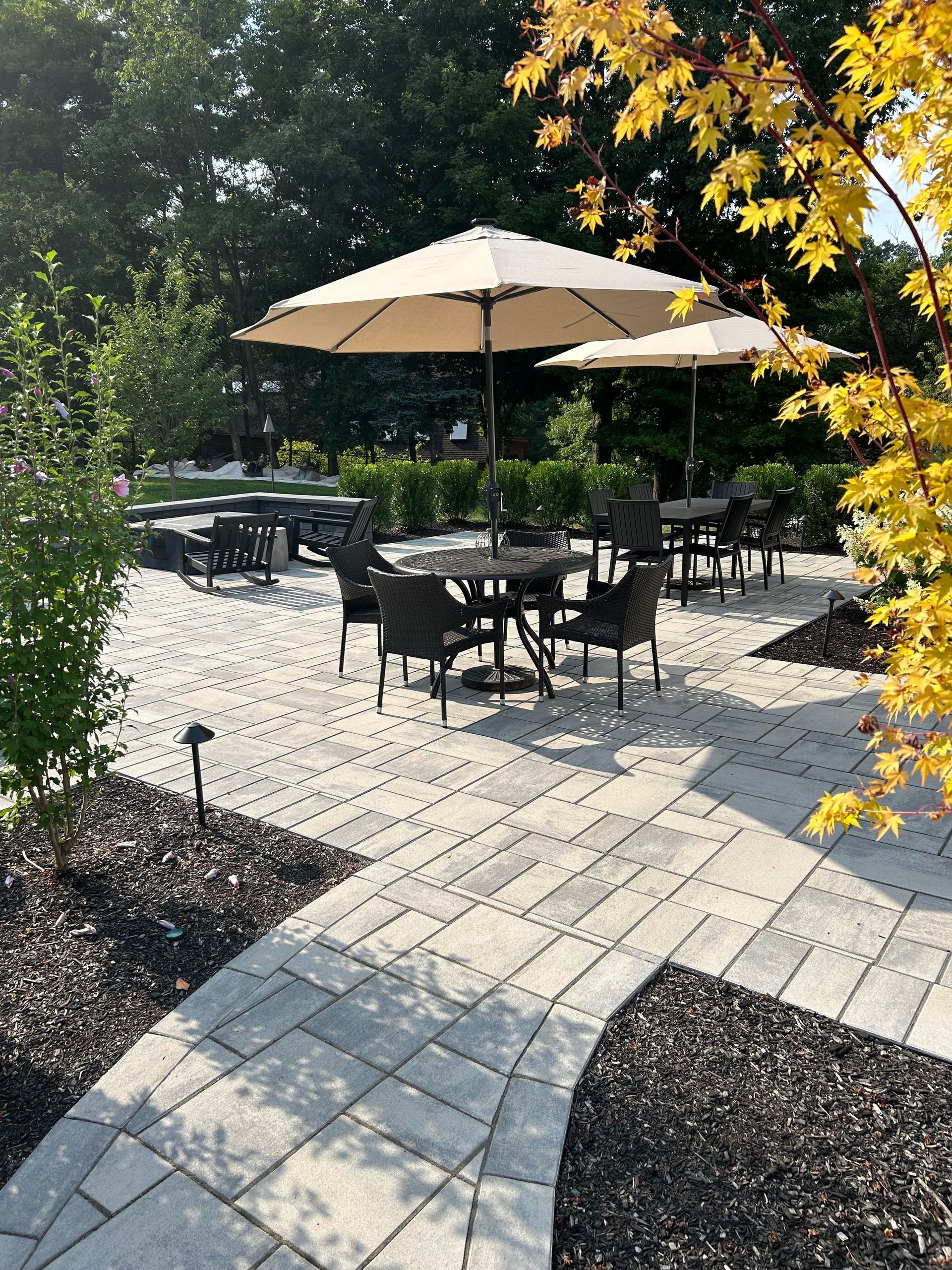 Outdoor patio with black tables and chairs under a beige umbrella, bordered by brick pavers and autumn foliage