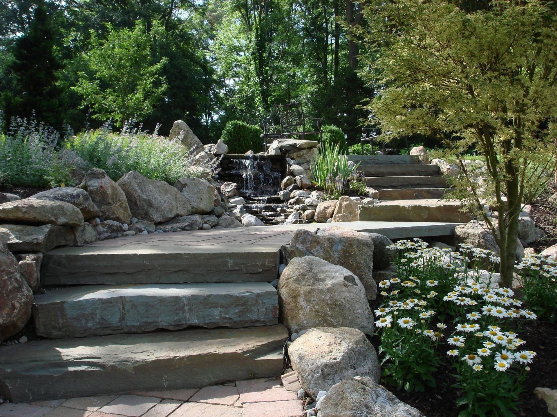 Stone steps in a lush garden leading to a small waterfall, with flowers and greenery around them