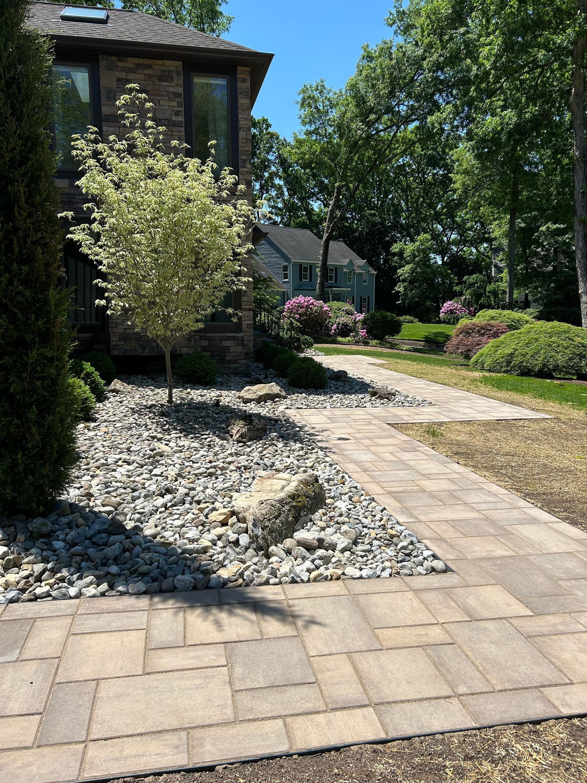 Stone walkway through a landscaped garden beside a house, with trees, shrubs, and bright sunlight