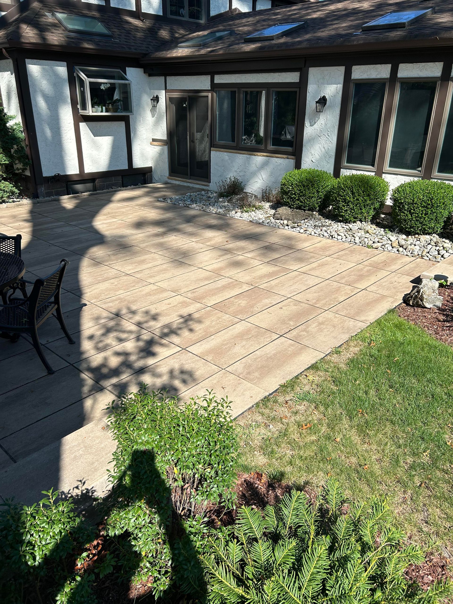 Sunlit stone patio outside a house, bordered by grass and shrubs, with a shadowed camera operator in the foreground