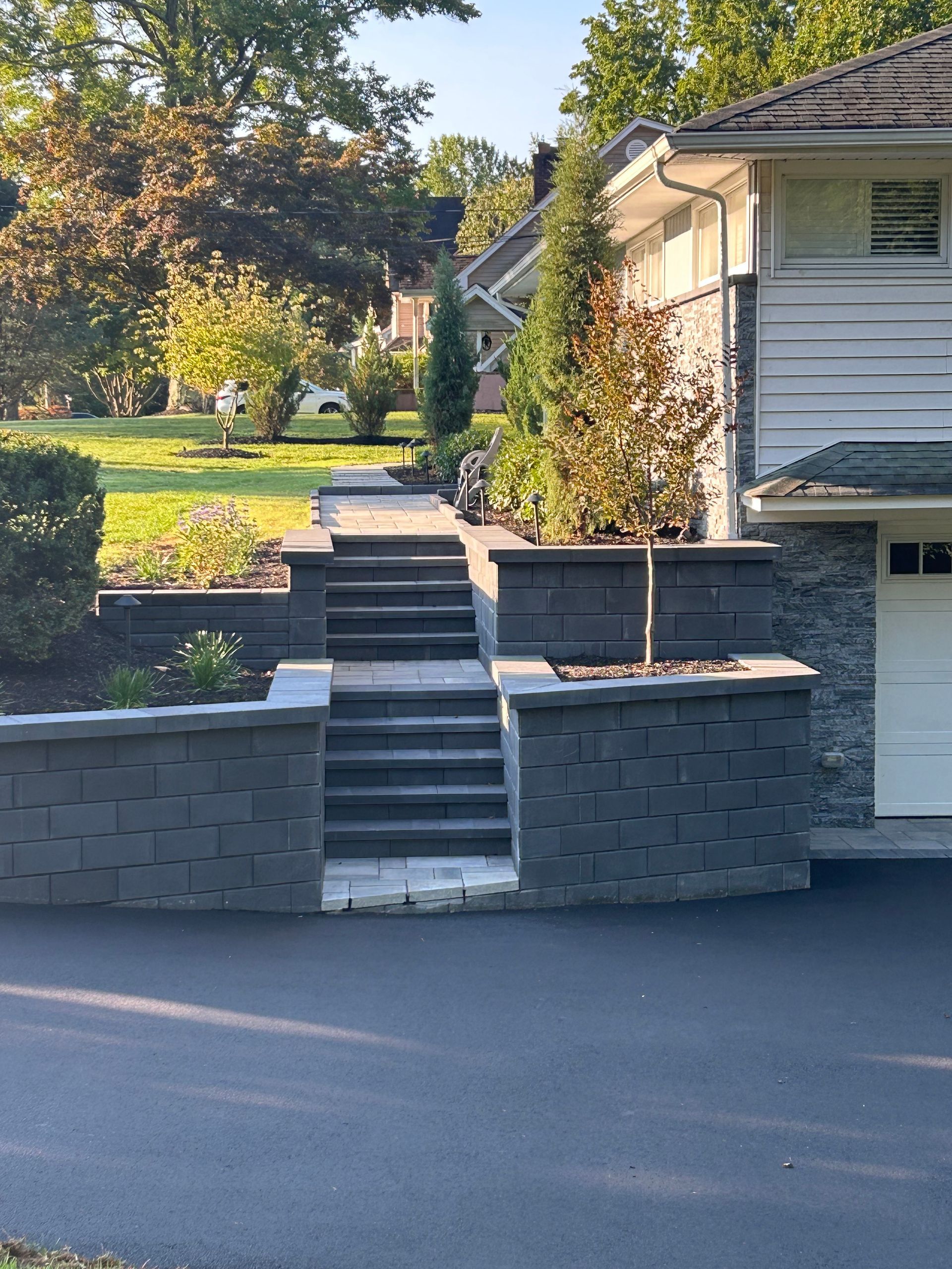 Stone stairway and retaining wall leading up to a sunlit front yard with a house and trees.