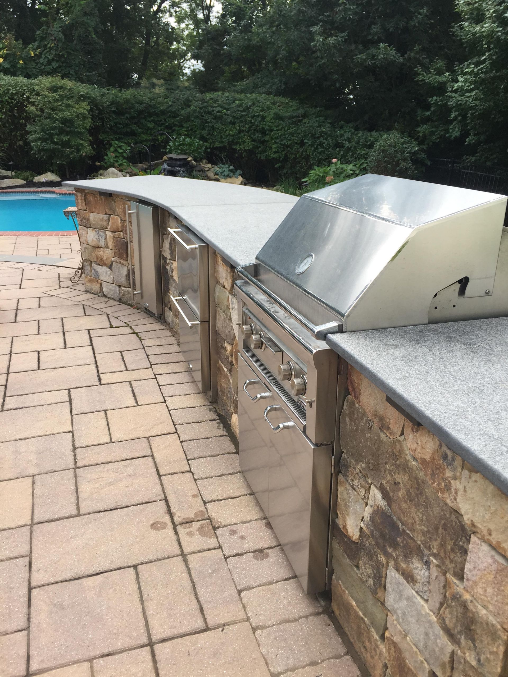 Curved stone poolside bench with metal cover beside a blue swimming pool and trees in the background