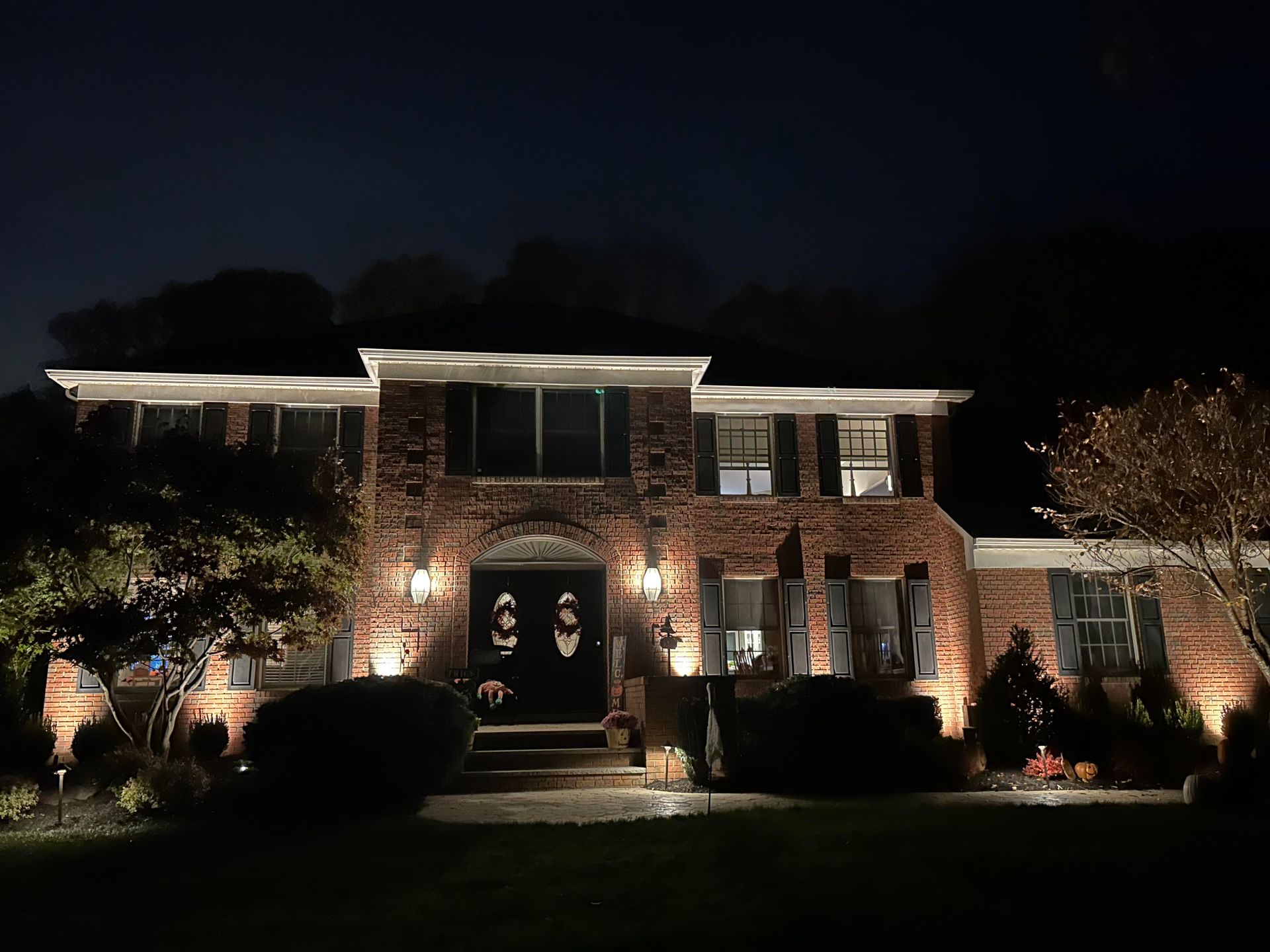 Illuminated brick house at night with lit windows, front steps, and landscaped yard under dark sky