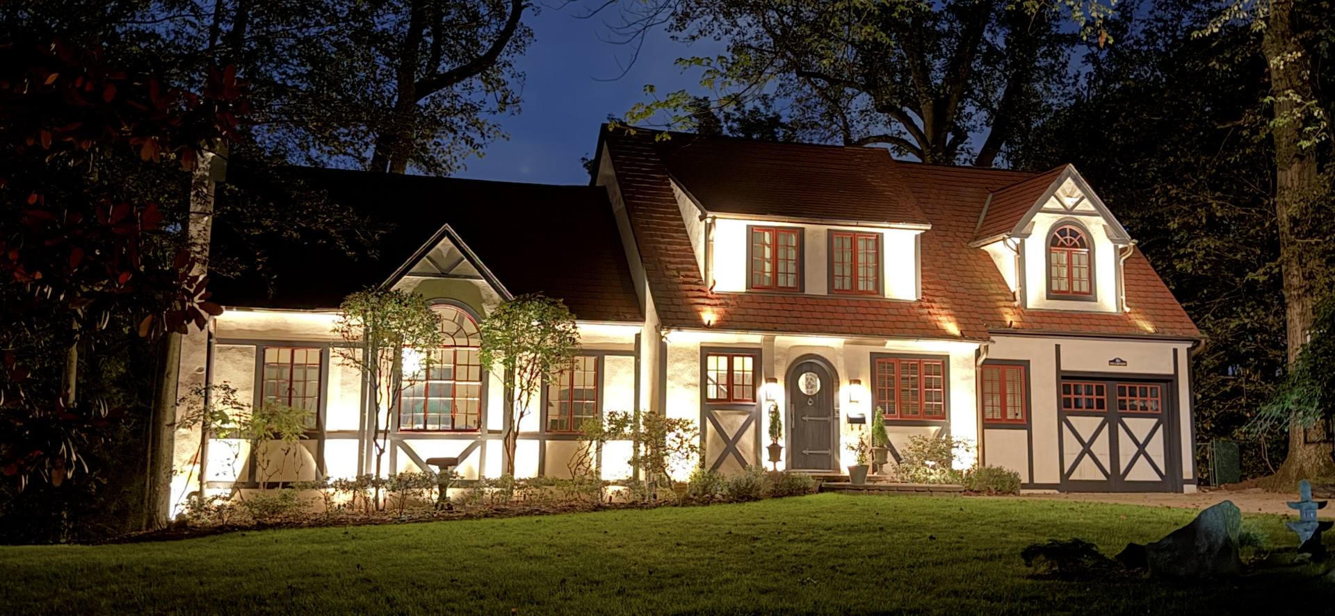 Illuminated two-story house at night with warm lights, white siding, and a garage, surrounded by trees.
