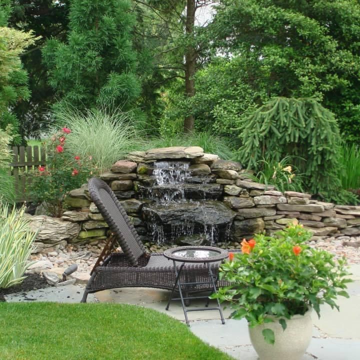 Backyard patio with wicker chair, small waterfall, lush greenery, and potted orange flowers