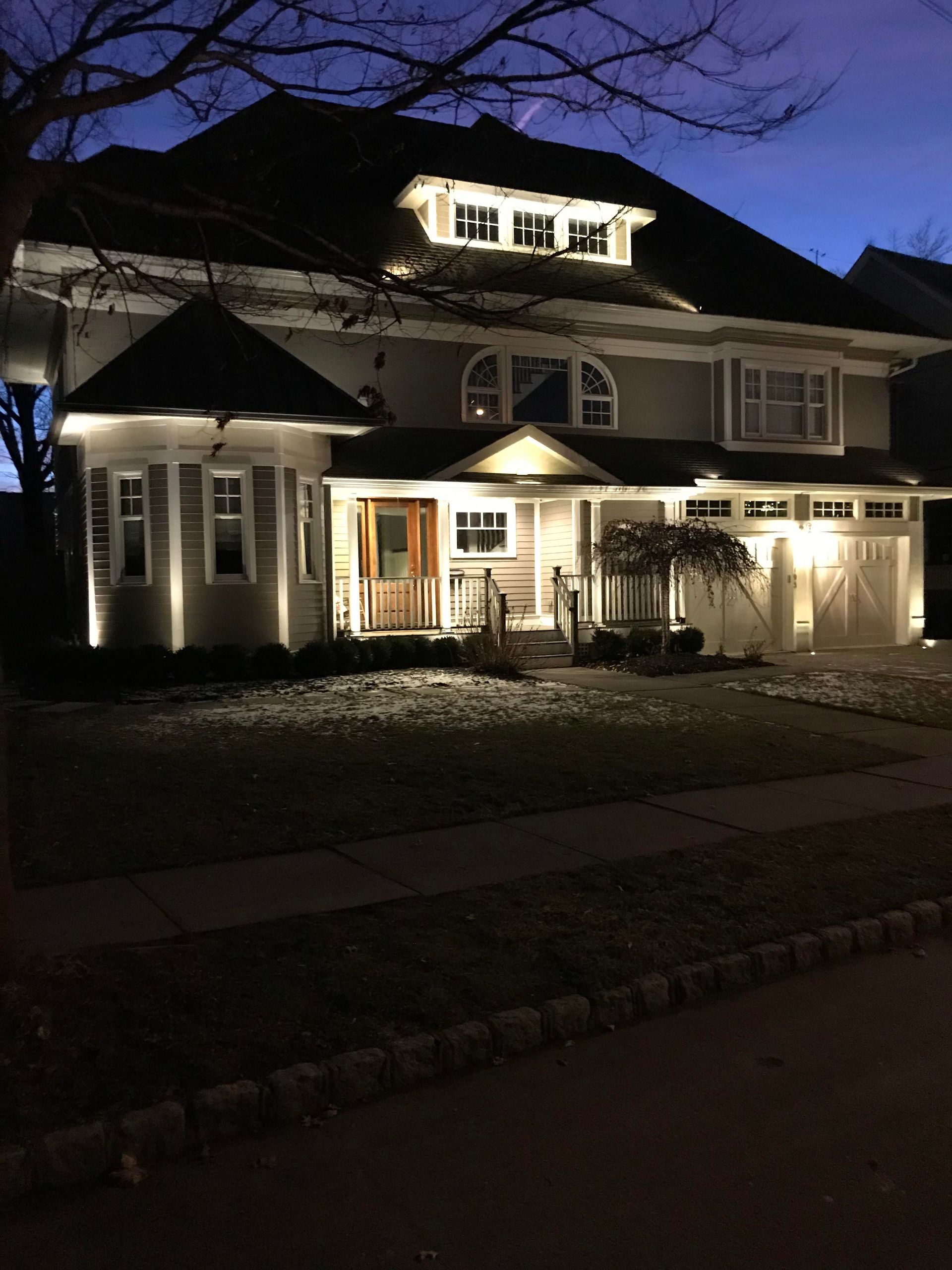 Illuminated two-story house at dusk with warm porch and window lights against a dark blue sky