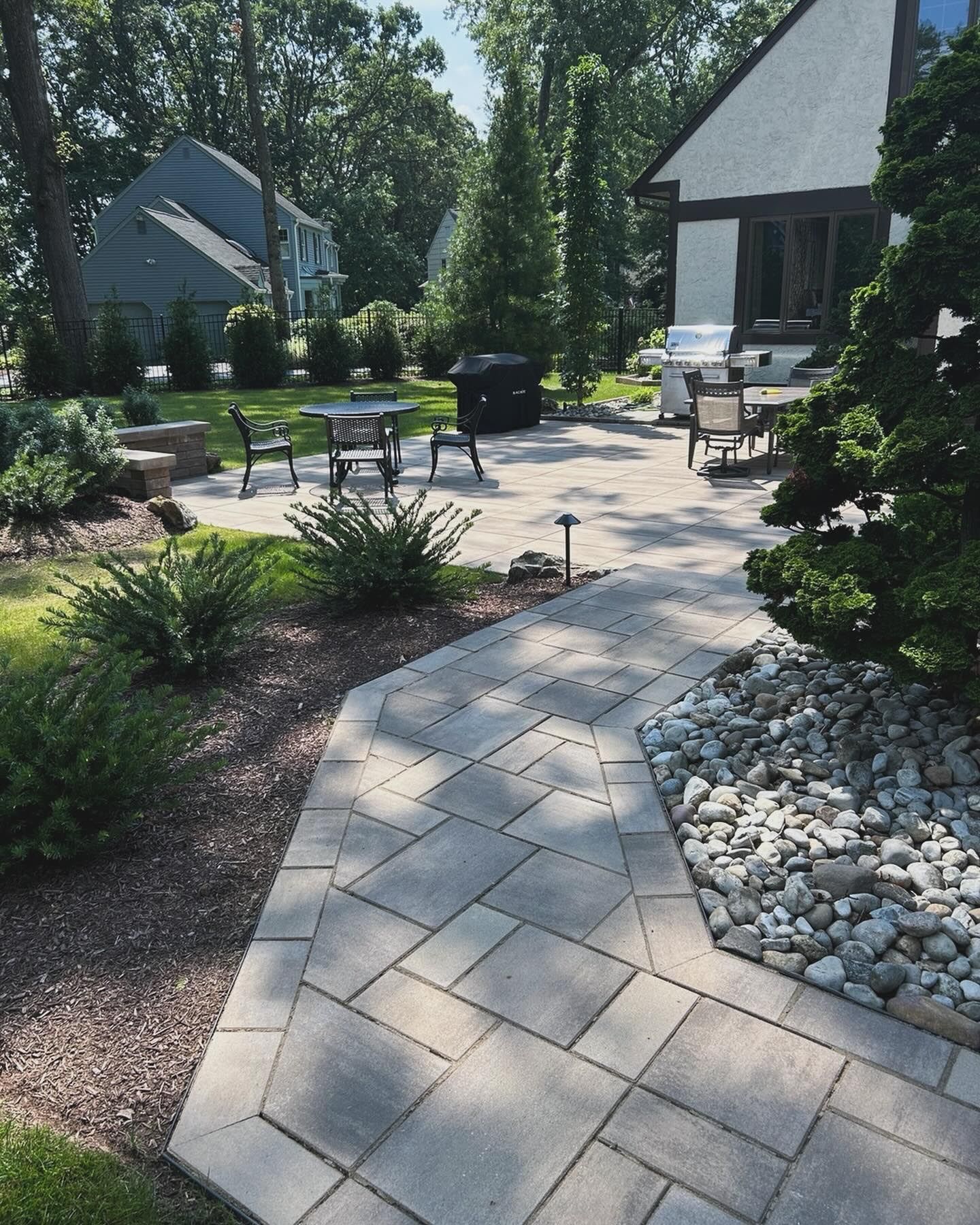 Modern landscaped patio with gray paver path, outdoor chairs, grill, and white stone border in sunlight
