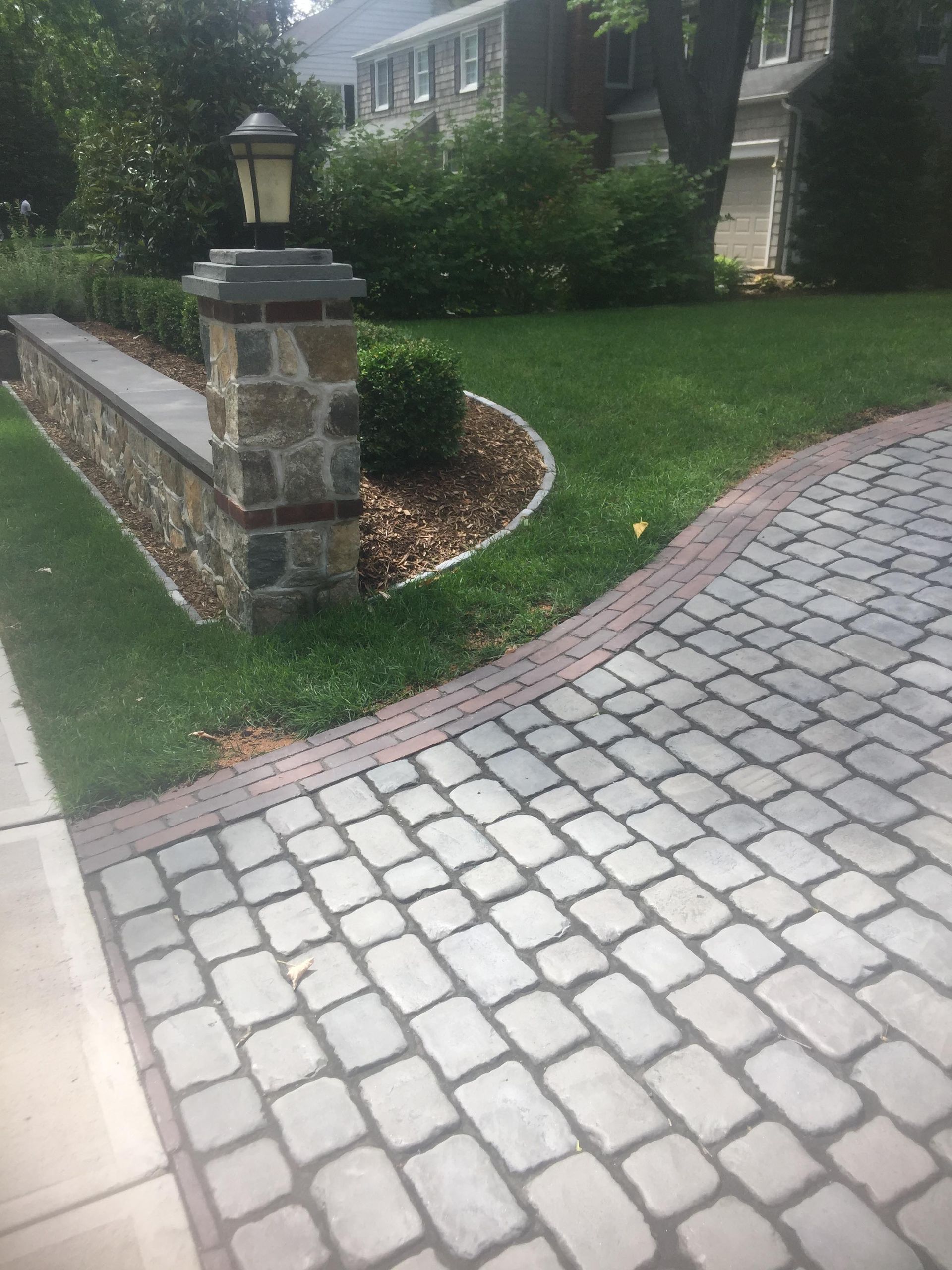 Brick-paver walkway beside a landscaped lawn and stone wall in front of a house