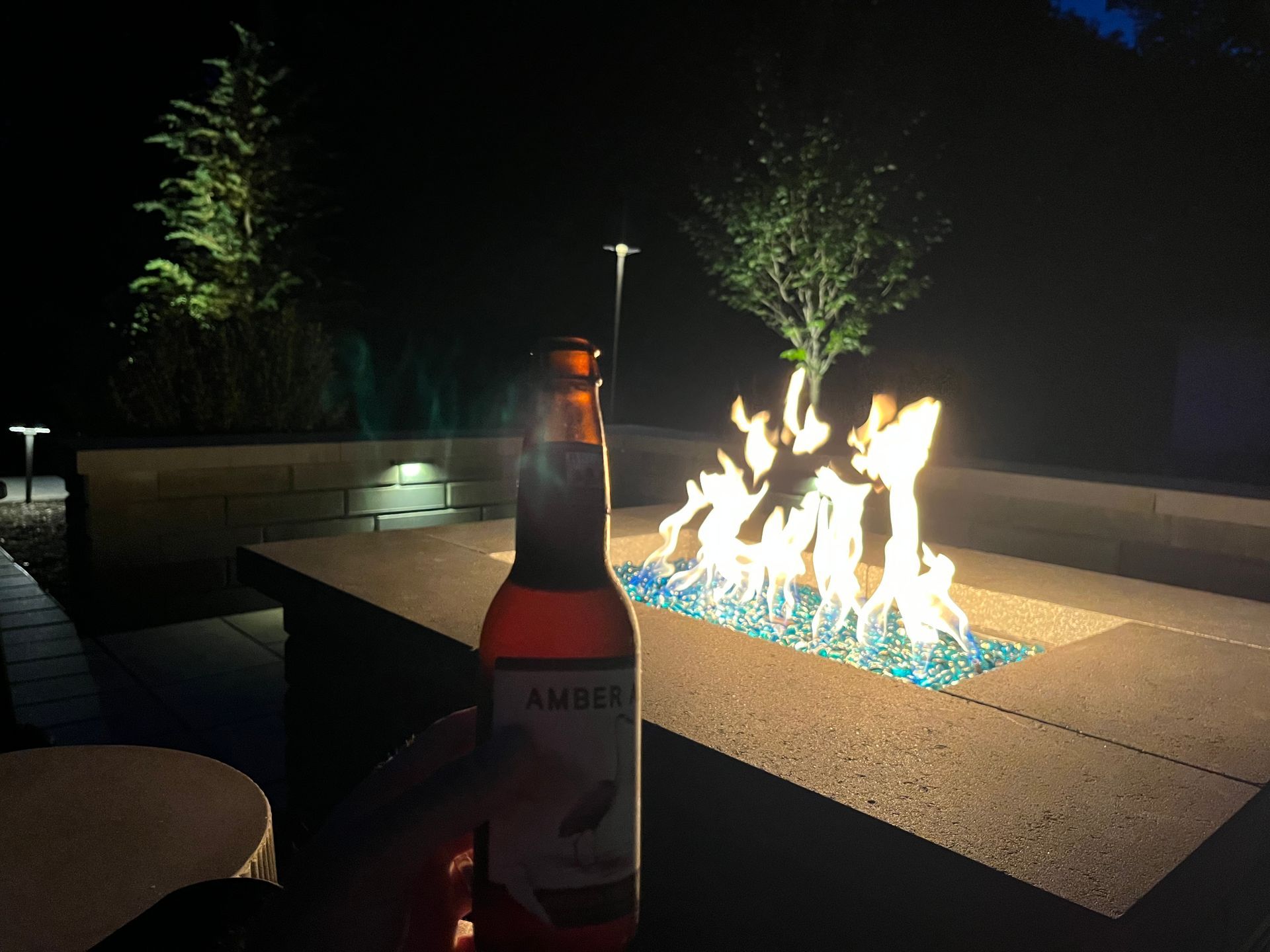 Bottle in foreground beside a fire pit with blue flames at night on a patio