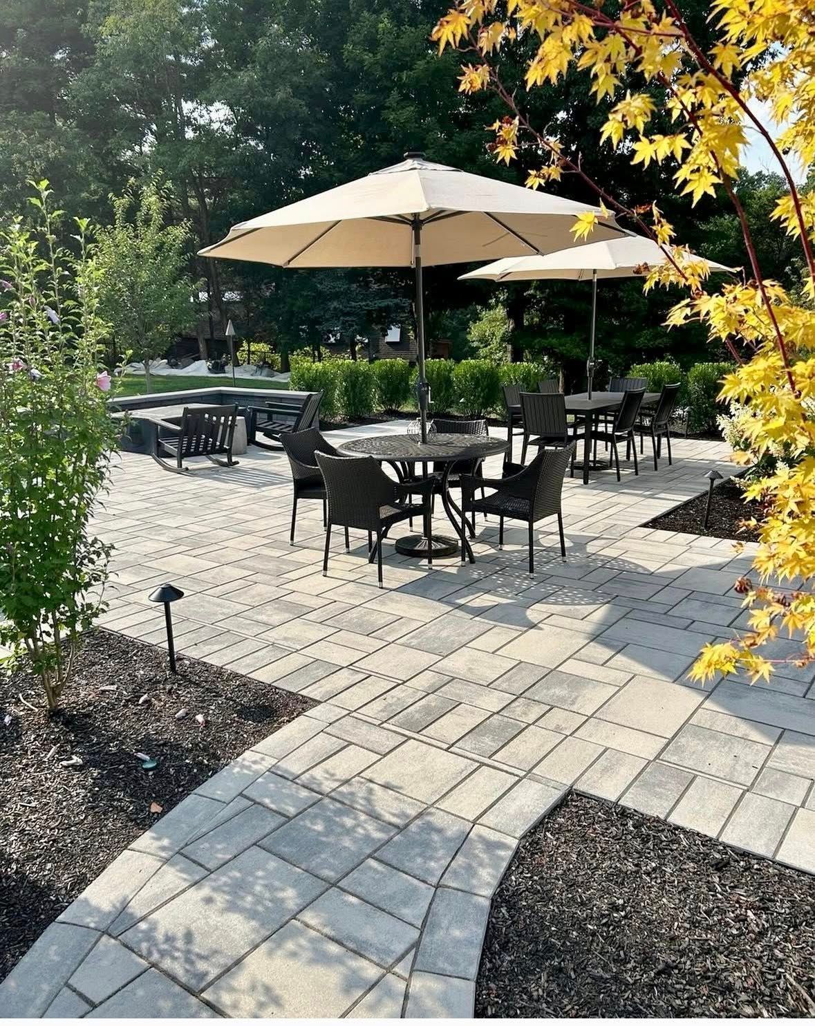 Outdoor patio with black tables and beige umbrellas, surrounded by greenery and a stone walkway.