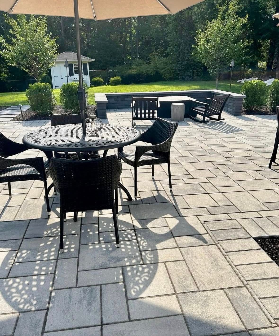 Patio with round table and black chairs on a sunlit stone terrace, with umbrella and benches nearby