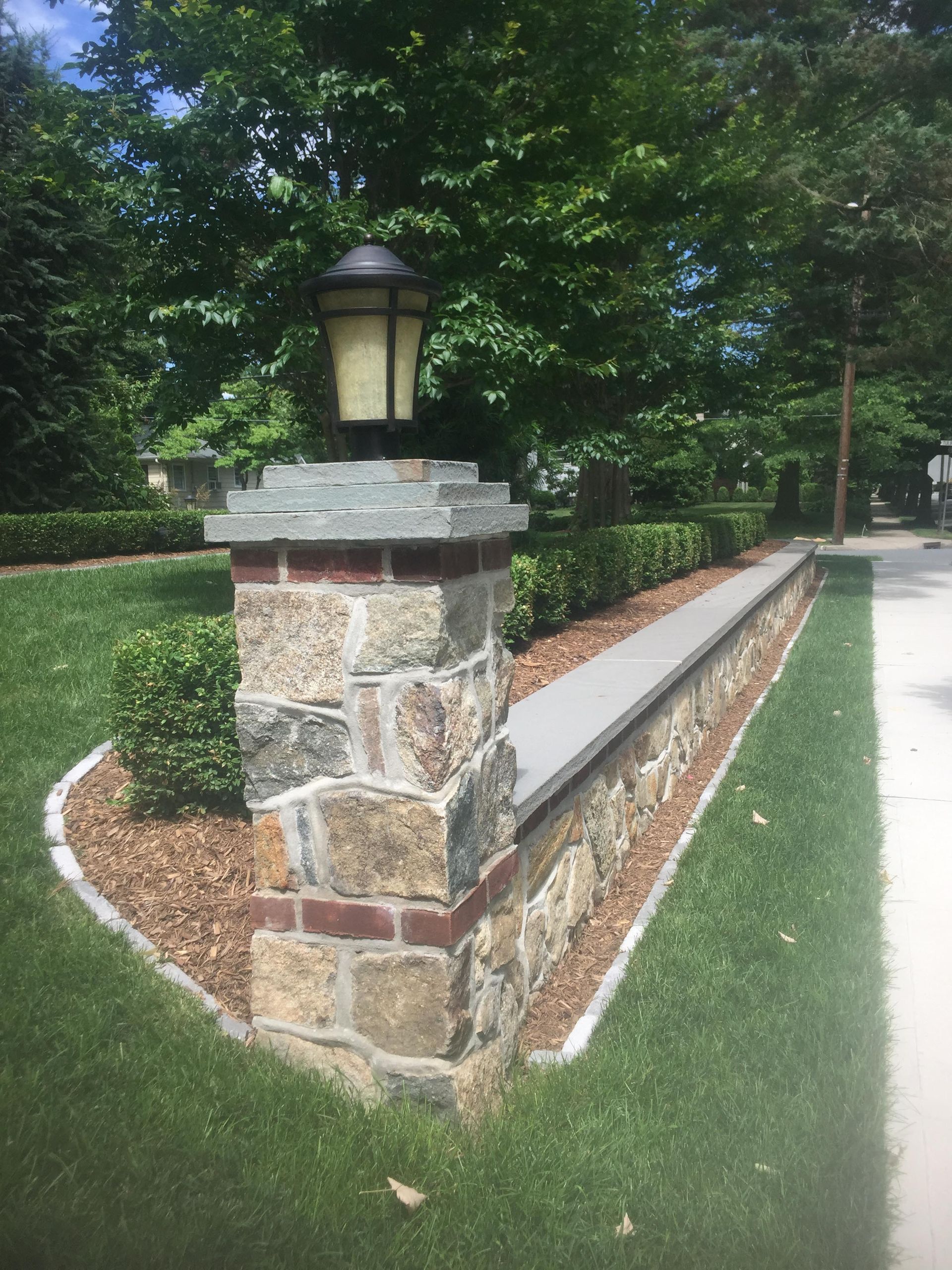 Stone pillar with lantern beside a low brick-and-stone wall along a manicured path in a leafy garden