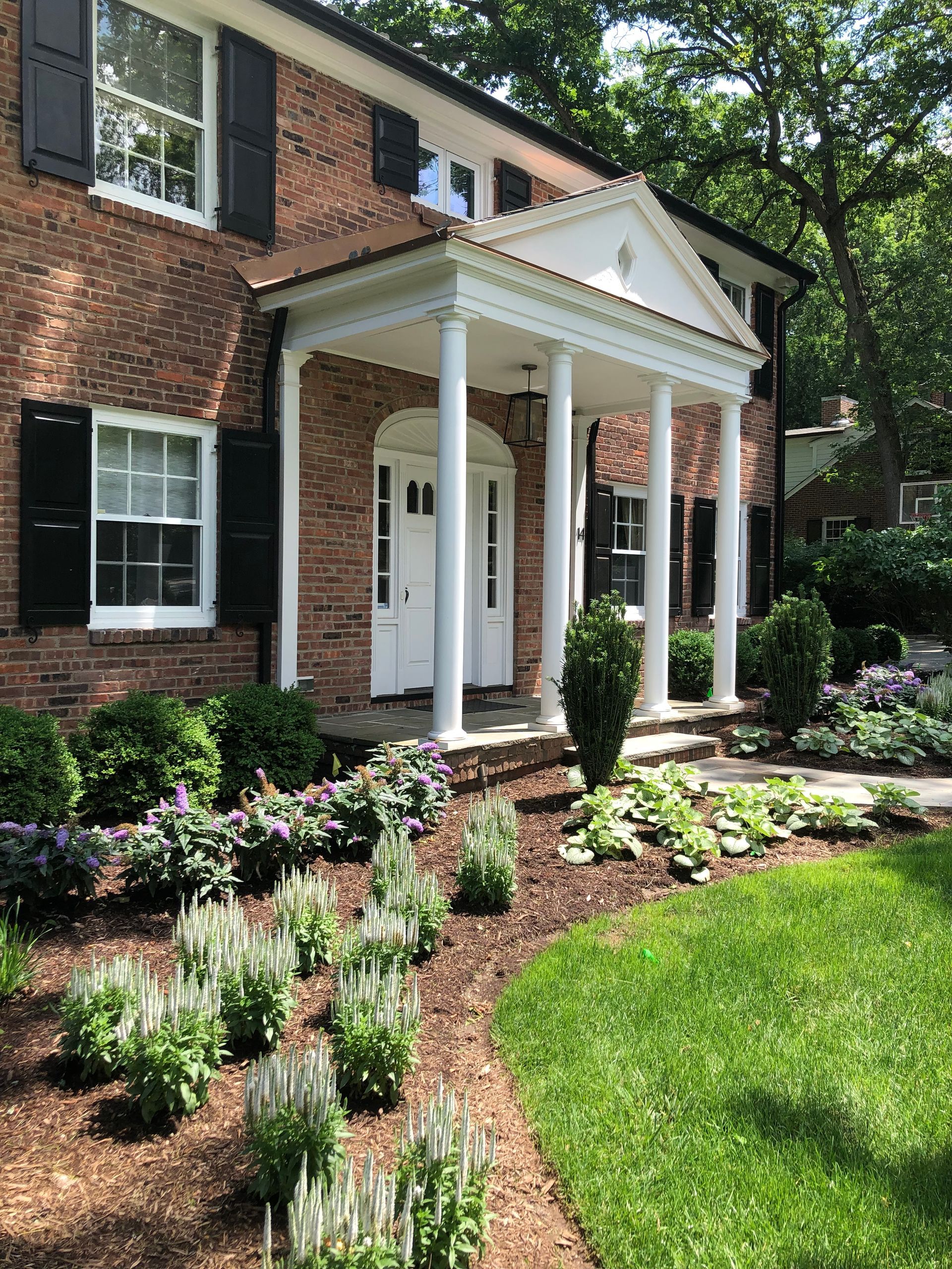 Brick house with white-columned front porch, landscaped garden, and green lawn in daylight
