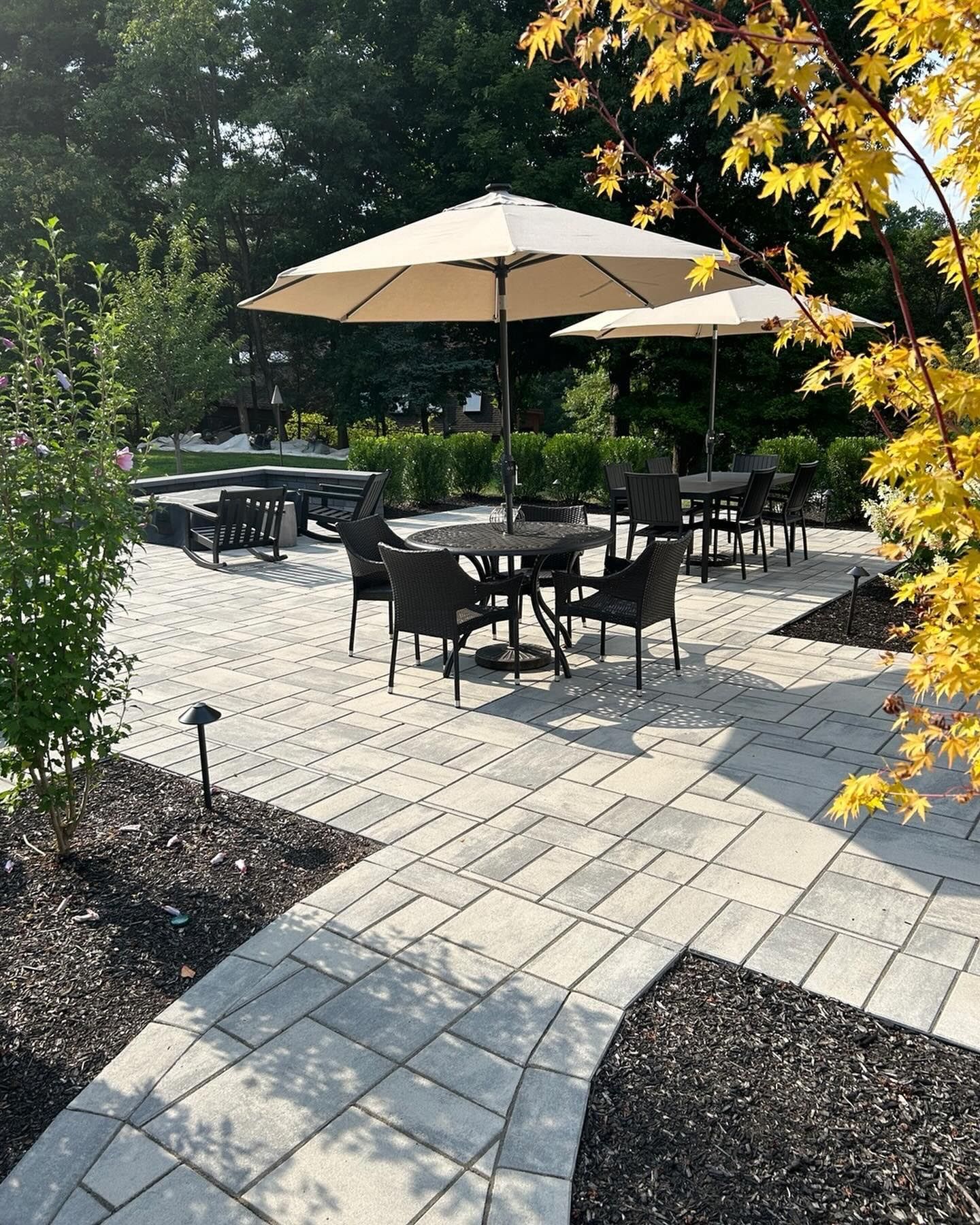 Outdoor patio with tables, beige umbrellas, and brick paving surrounded by garden beds and trees
