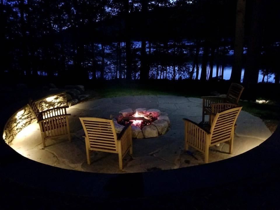 Outdoor fire pit seating area at night with three wooden chairs and a small lantern-lit table under trees