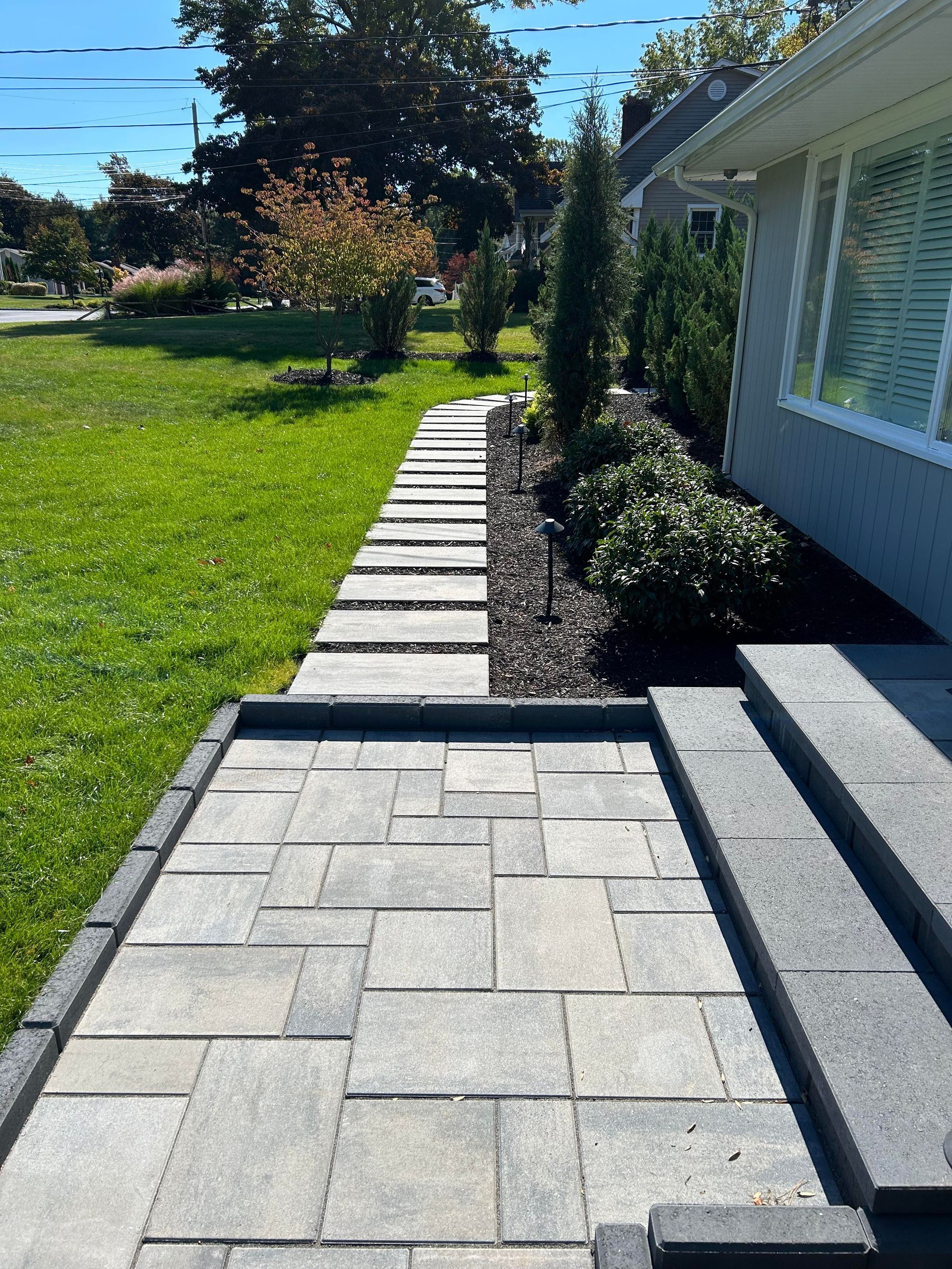 Paver walkway beside a house with steps, bordered by grass and shrubs in a sunny yard