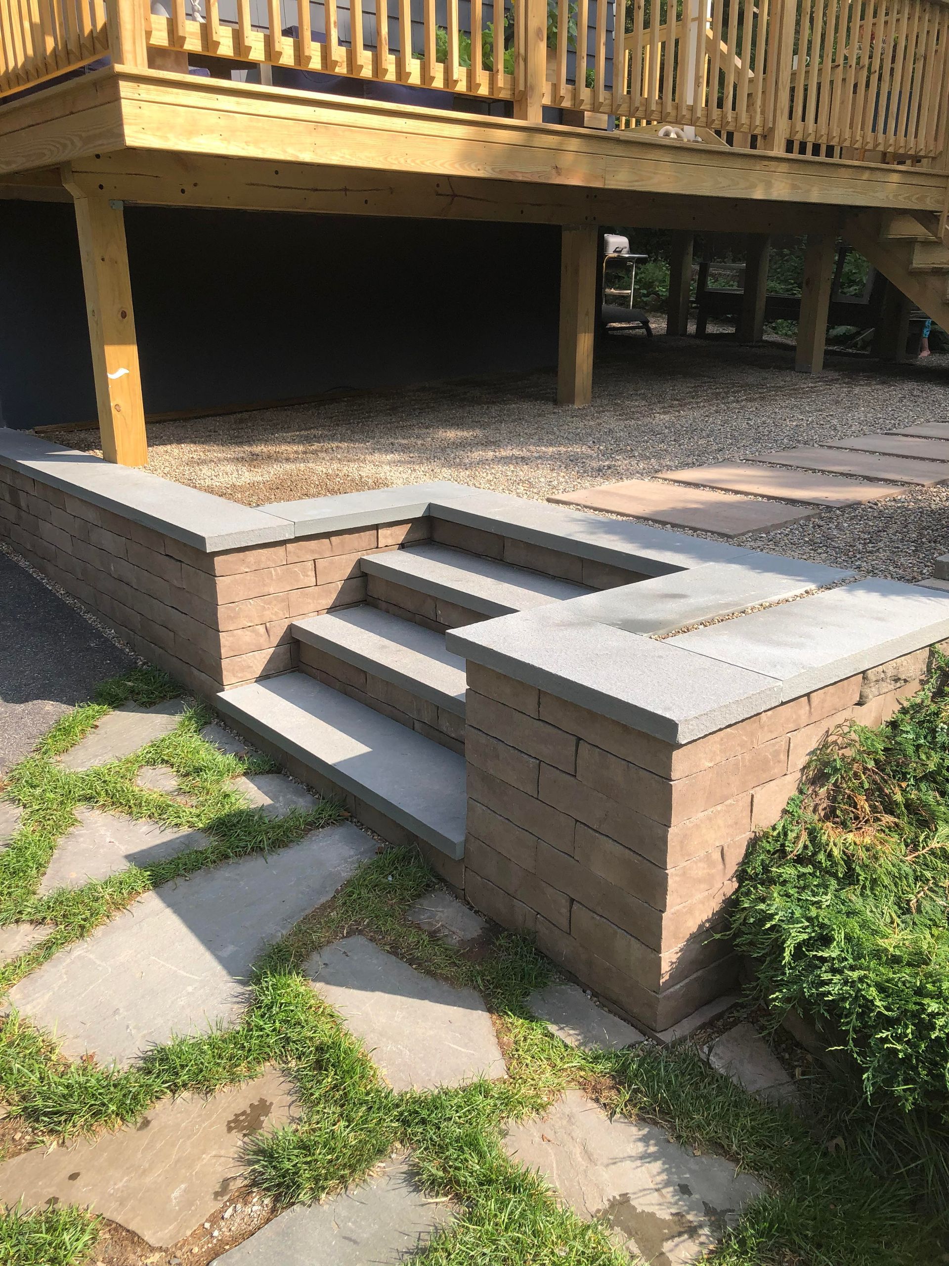 Raised brick patio steps and built-in benches beneath a wooden deck, with stone pavers and grass nearby.